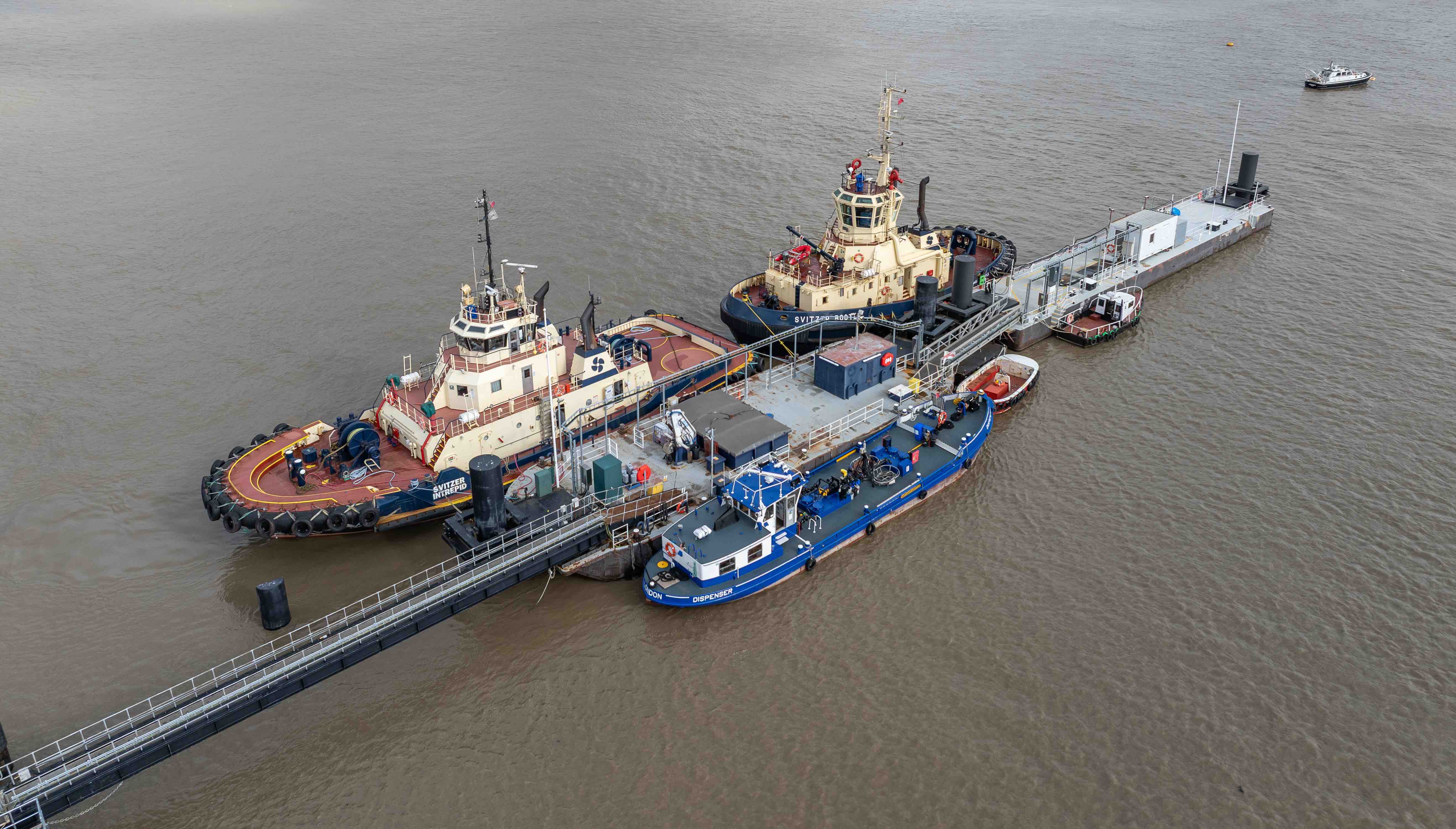 Three tugboats docked alongside a long platform structure in murky brown water.