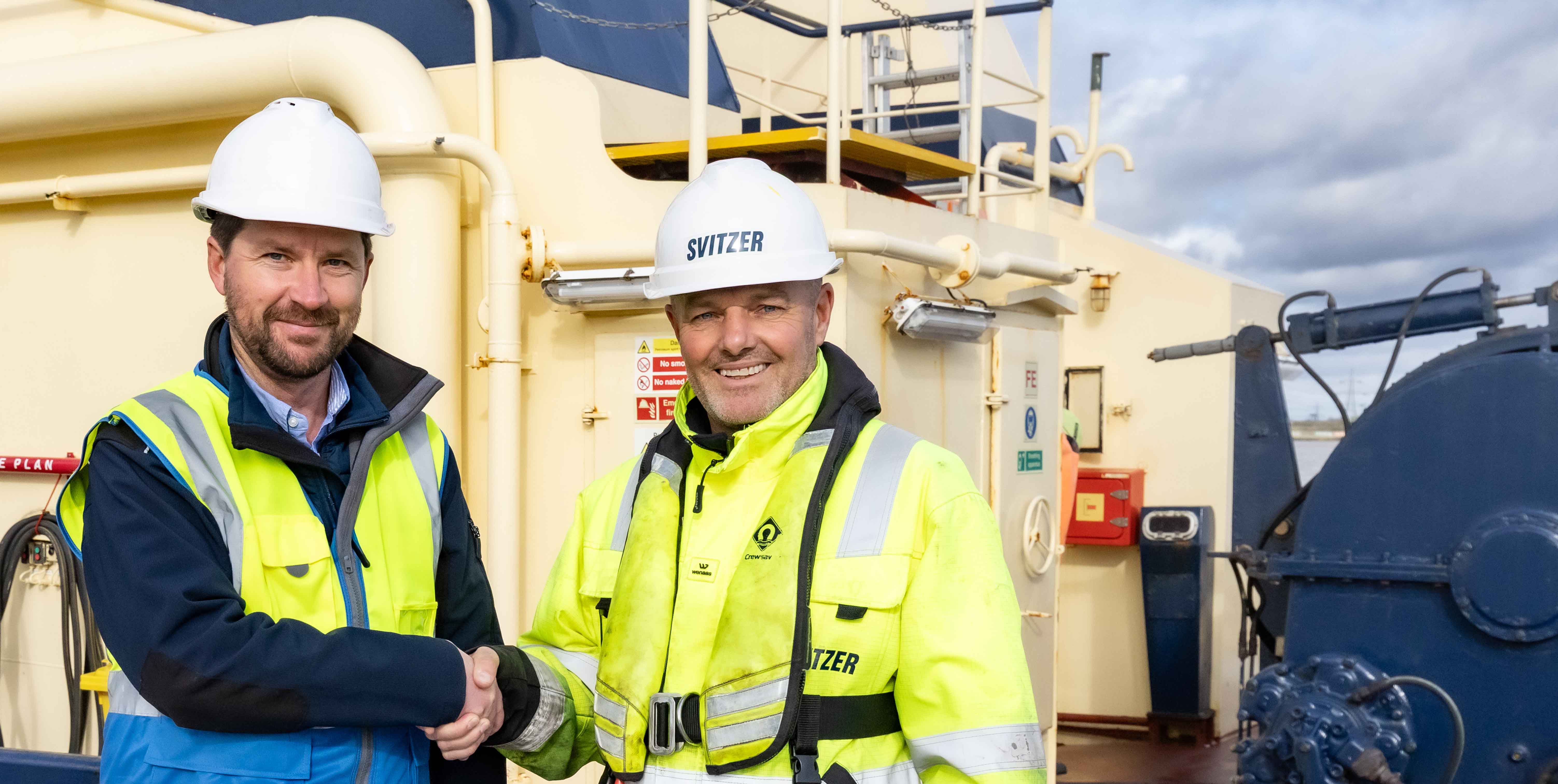 Two men in safety gear and hard hats shaking hands on a ship deck.