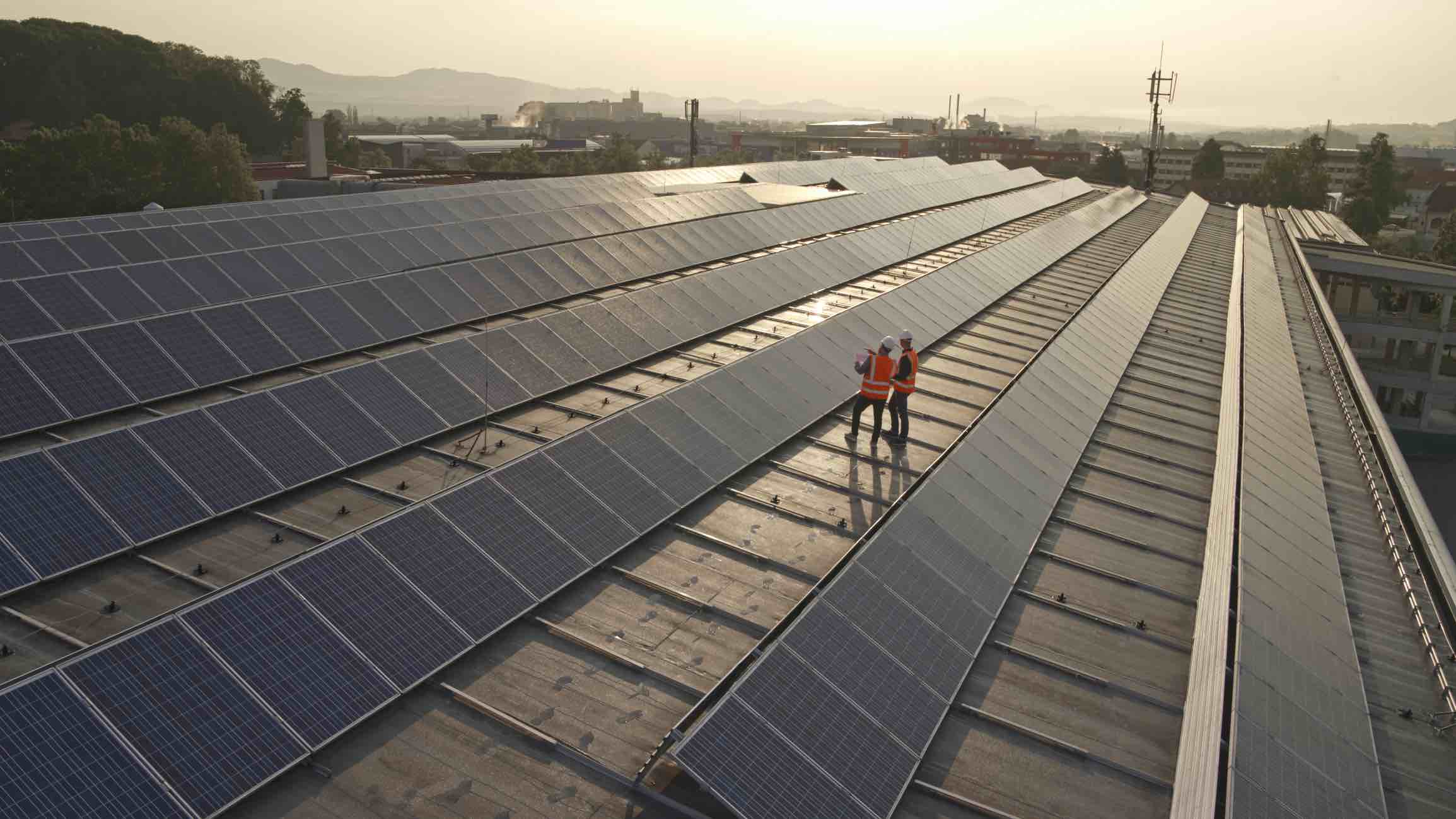 Two workers in safety vests inspecting solar panels on a large rooftop at sunset.
