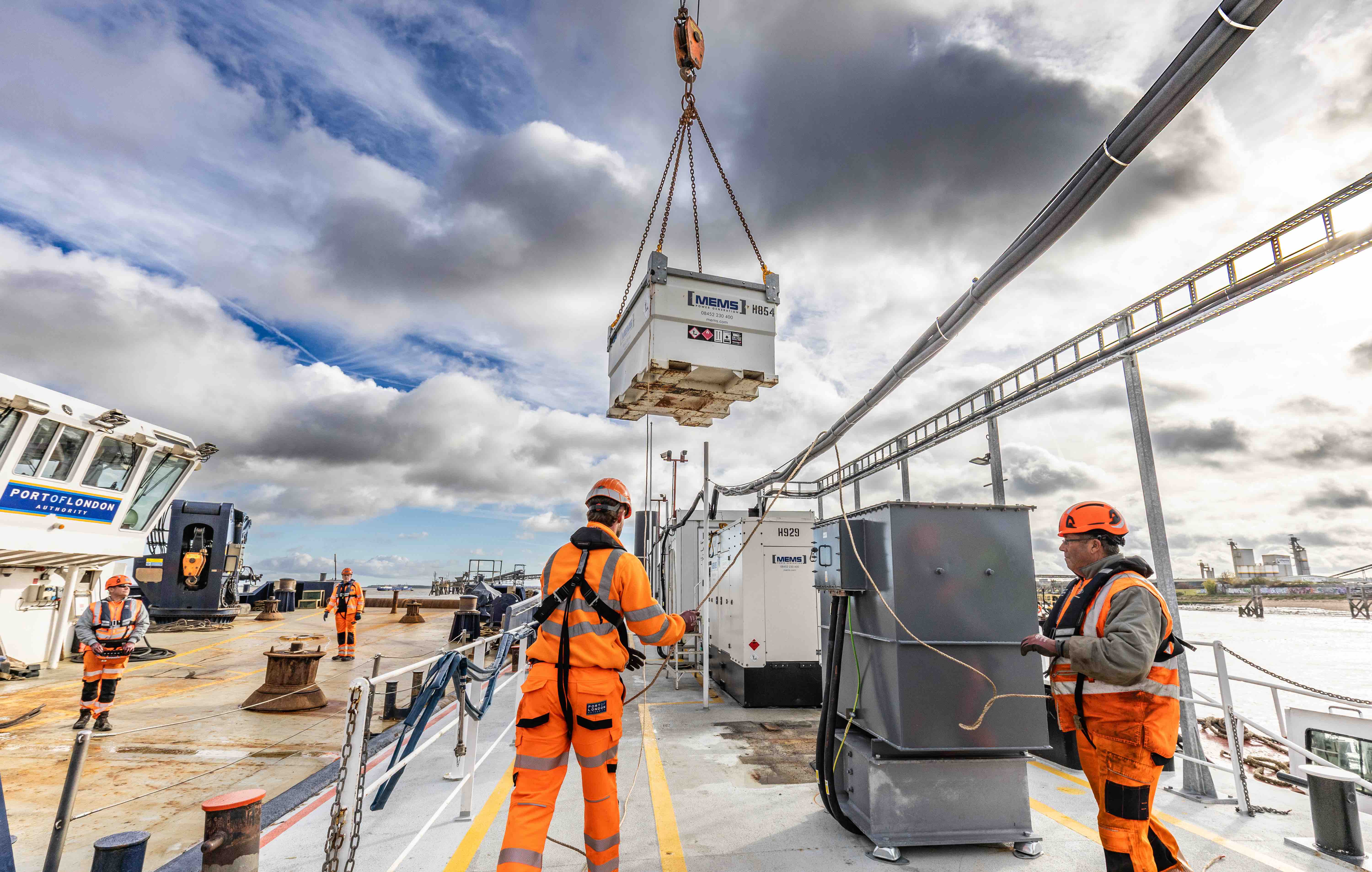 Workers in orange safety gear guide a suspended container on a ship deck under a partly cloudy sky.
