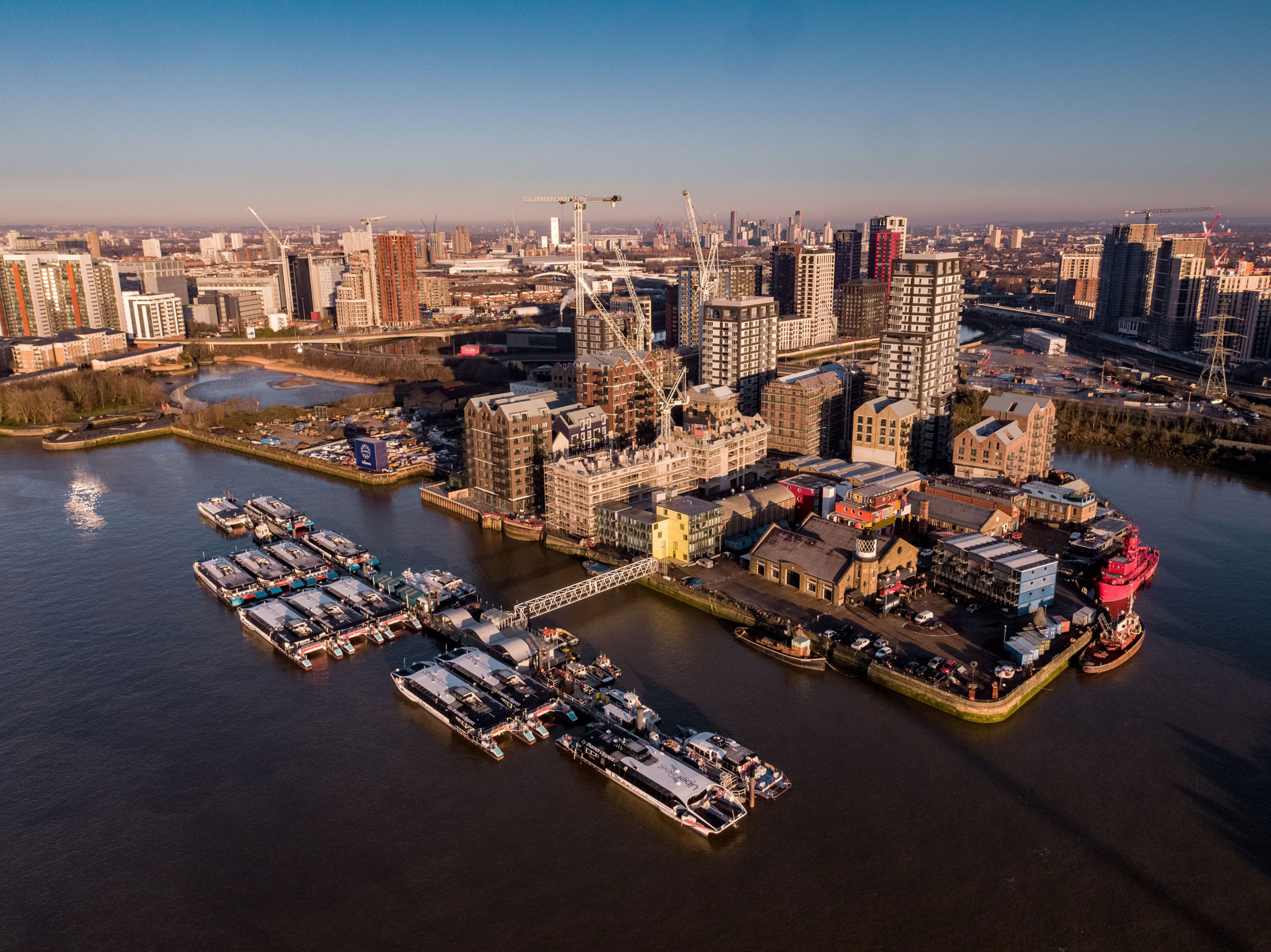 Aerial view of a riverside urban area with boats docked, buildings, cranes, and a city skyline in the distance.