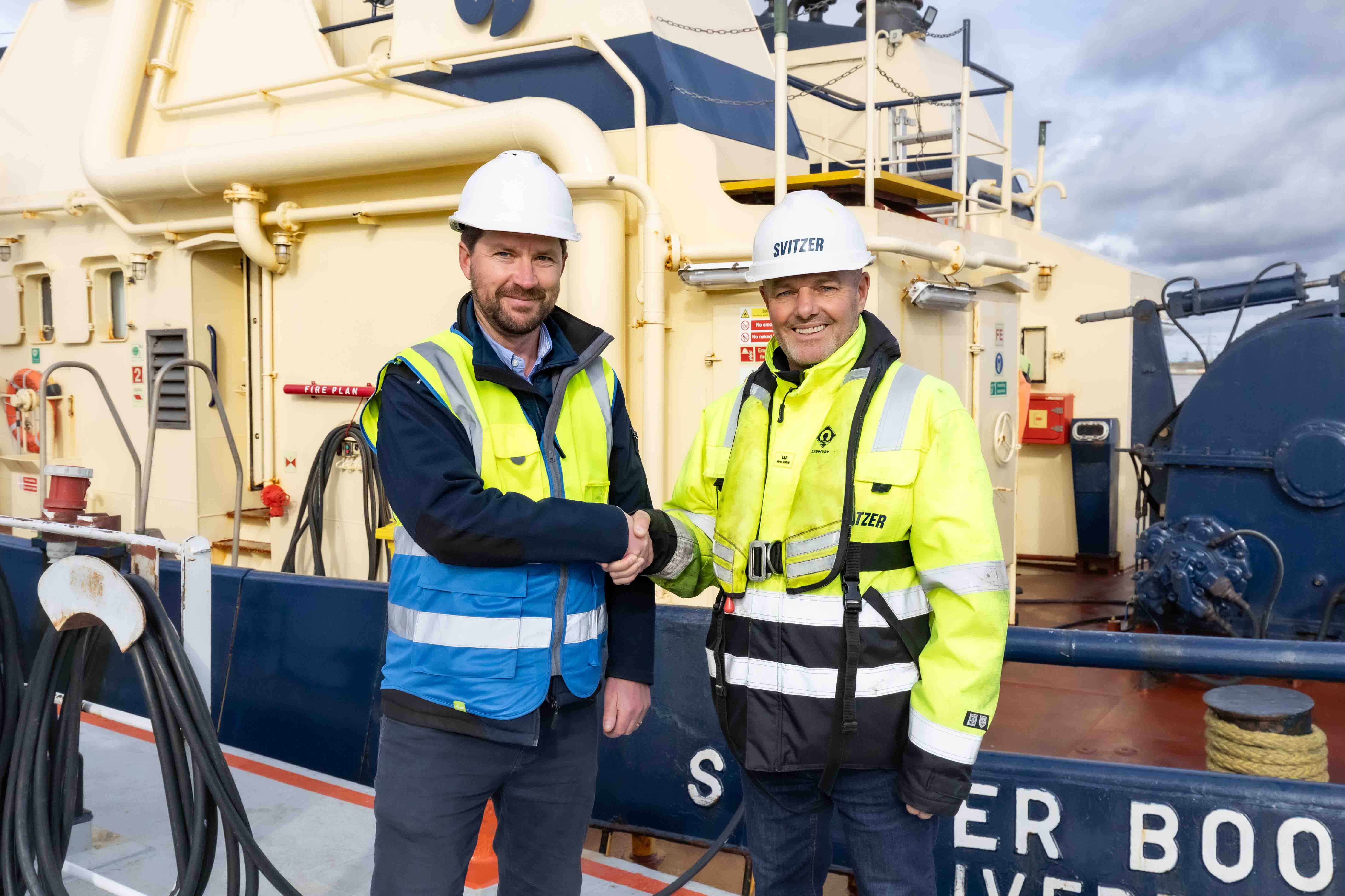 Two men in hard hats and safety vests shaking hands on a ship deck with industrial equipment.