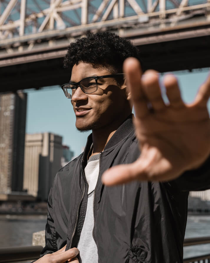 Young man wearing glasses and a black jacket reaching out a hand towards the camera with a bridge and city buildings in the background.