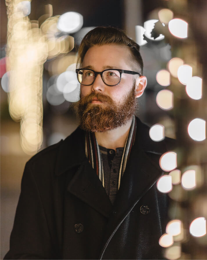Bearded man with glasses wearing a black coat and scarf, looking to the side with blurred decorative lights in the background.