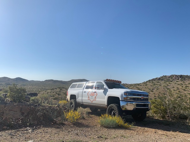 White off-road truck with logo parked on desert terrain with hills and clear blue sky.