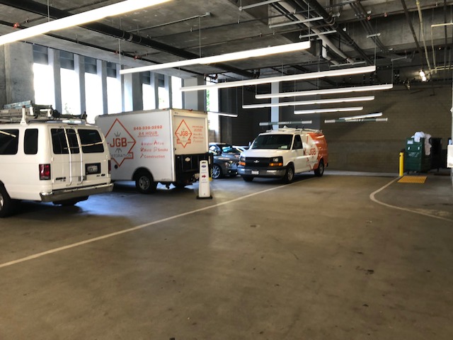 Indoor parking area with work vans, including two JGB Total Services vans and a black sedan, under fluorescent ceiling lights.