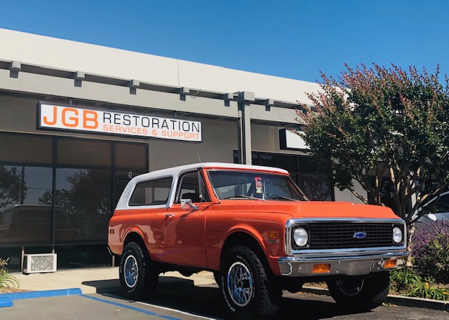 Orange vintage Chevrolet SUV with white roof parked in front of JGB Restoration Services & Support building under clear blue sky.