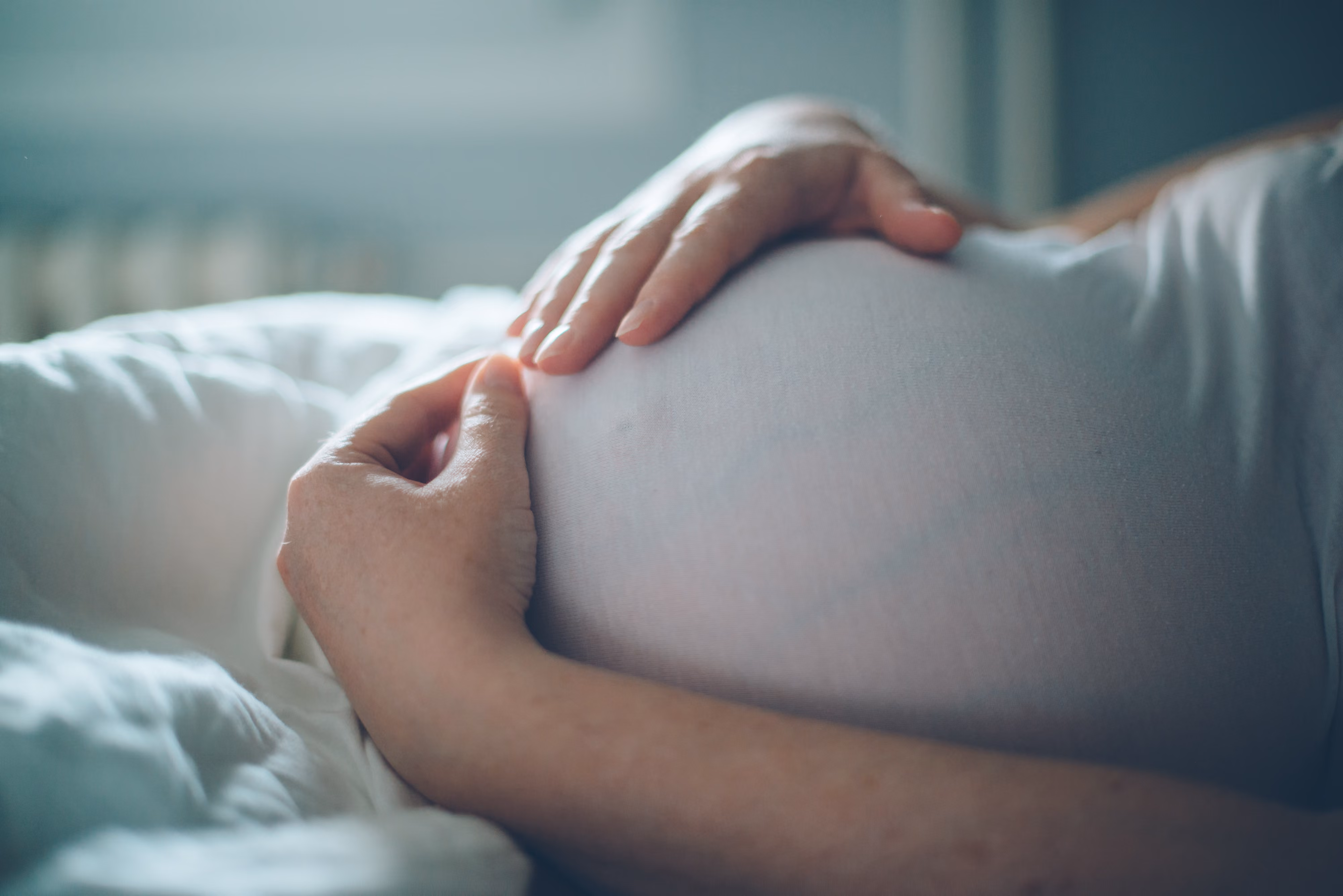 Person gently holding a pregnant belly while lying down with white bedding.