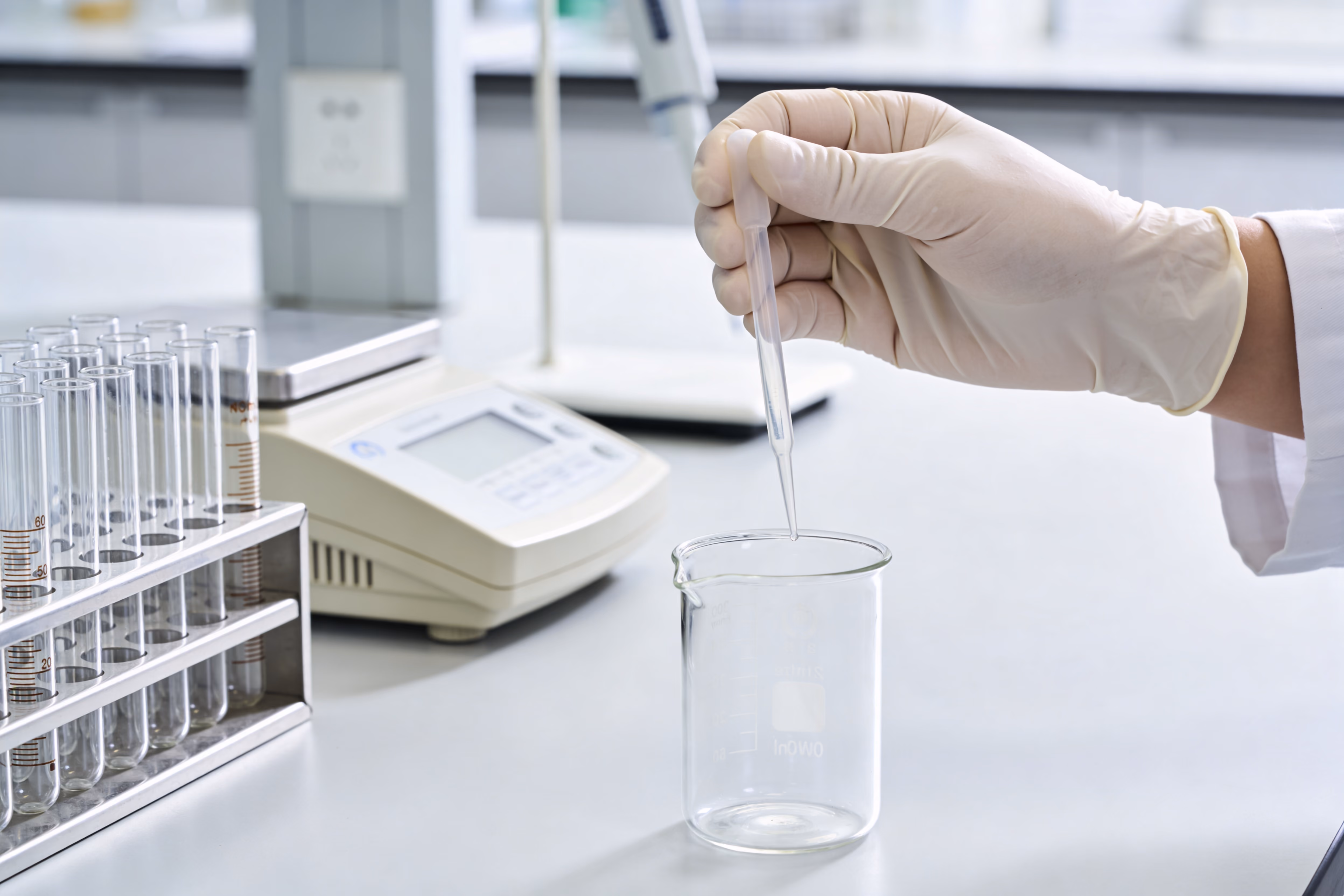 Gloved hand using a dropper to add liquid to a glass beaker in a lab setting.