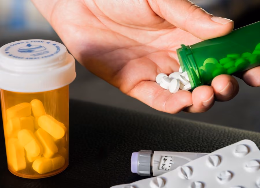 Hand pouring white pills from a green bottle with yellow capsules and medication blister packs nearby.