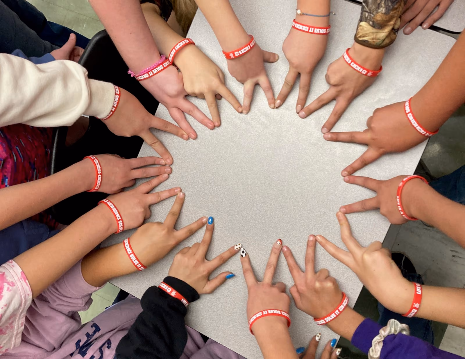 Circle of hands wearing red bracelets showing peace signs on a gray table.