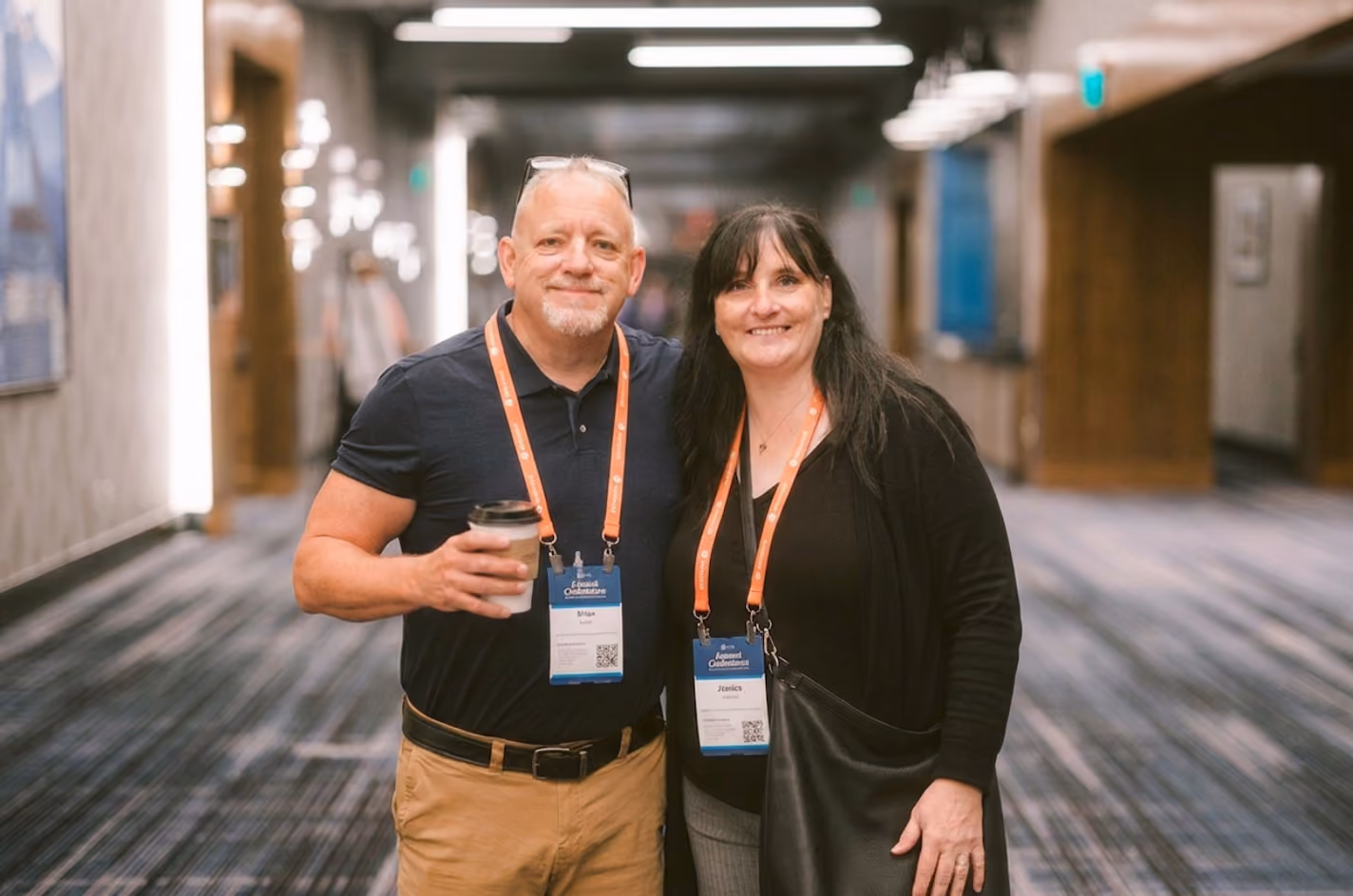 Two people with conference badges smiling and posing in a hallway, one holding a coffee cup.