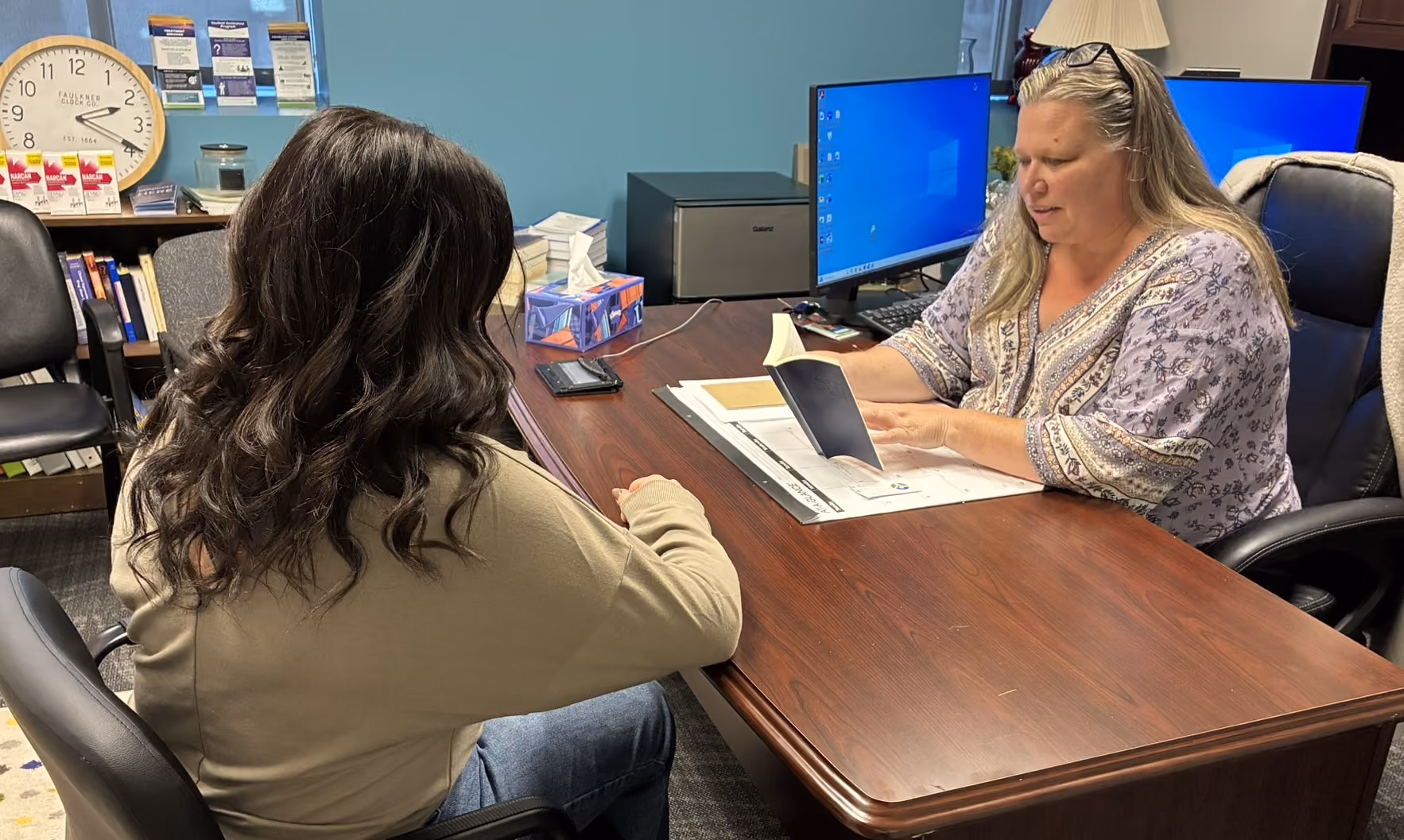 Two women seated across a desk in an office, one reviewing a booklet and the other listening.