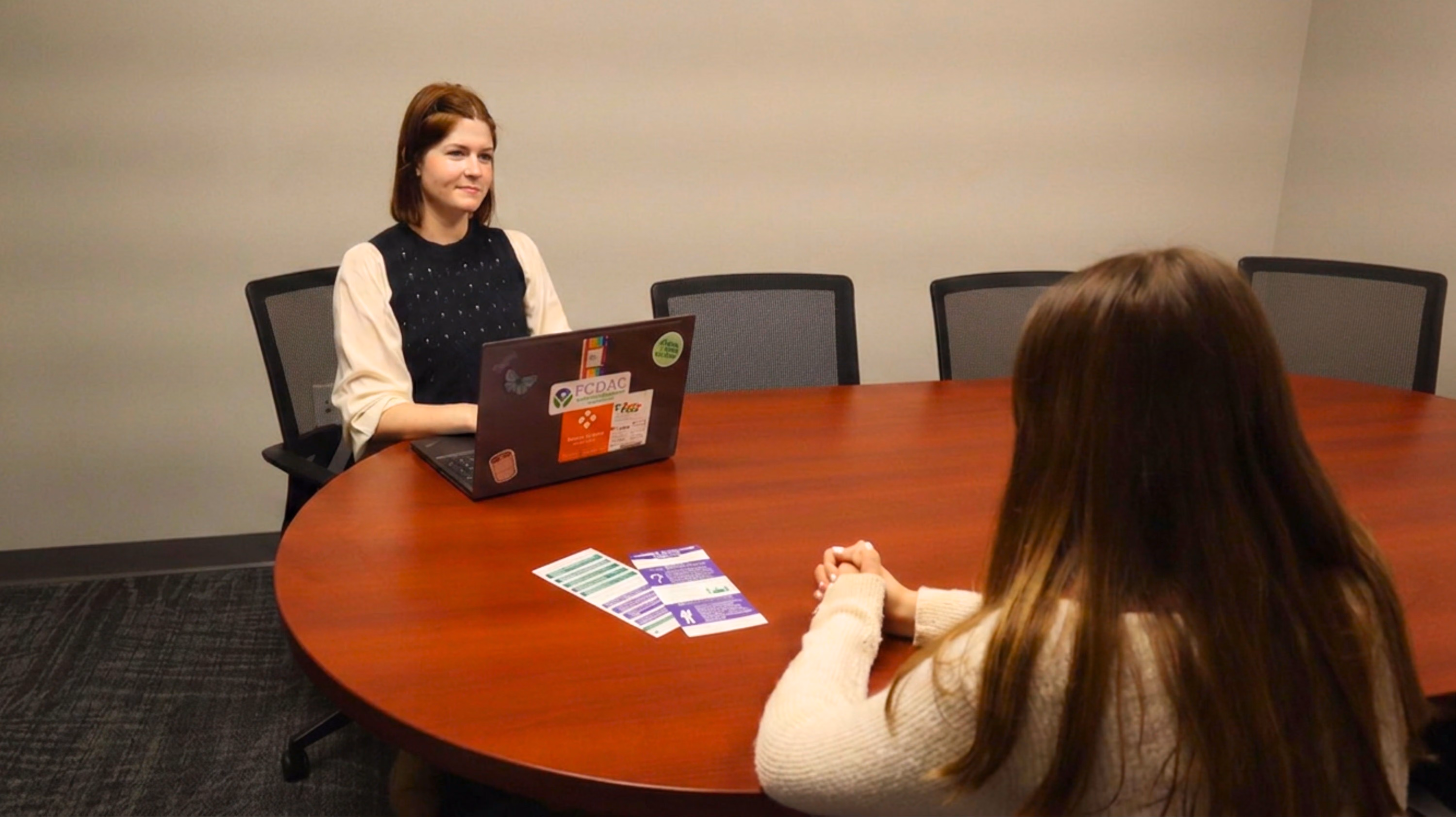 Two women sitting at a round conference table during a meeting, one with a laptop and papers.