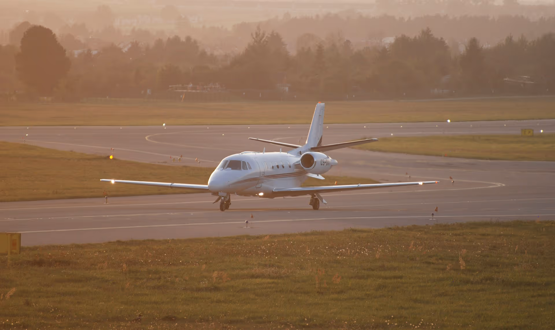 Small private jet taxiing on runway at sunset with trees and misty background.