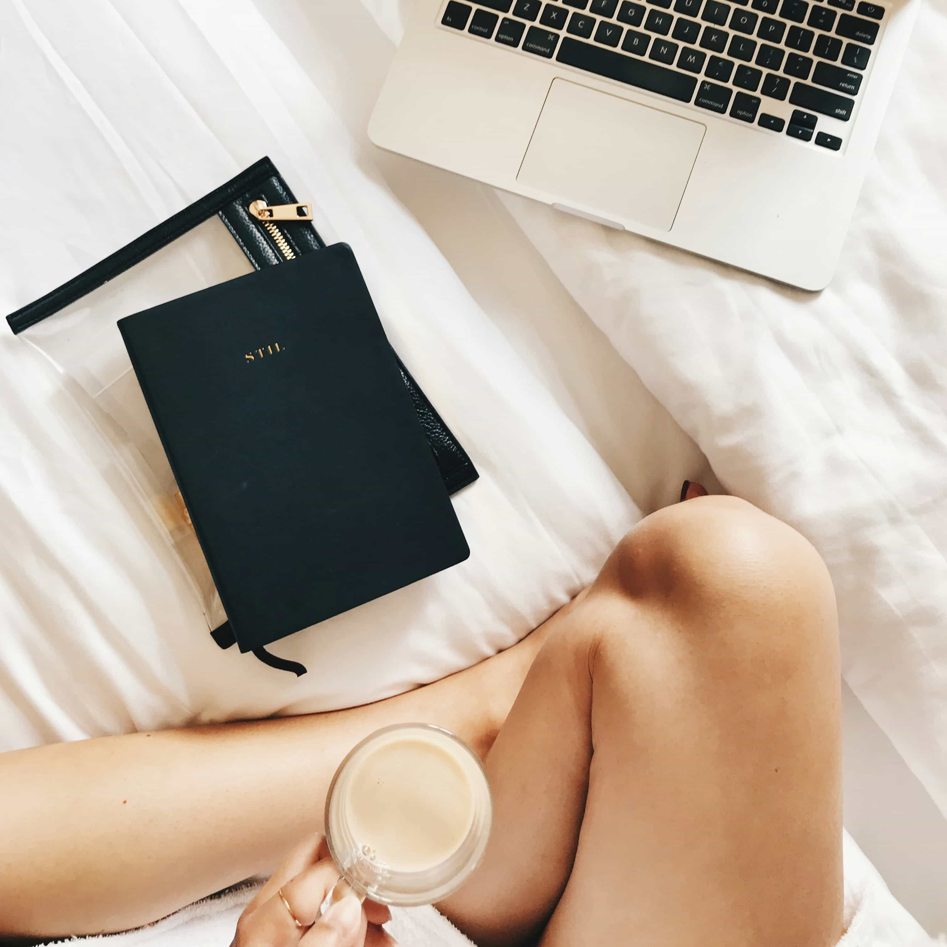 Overhead view of a person's crossed legs on a bed beside an open laptop, a black journal, and a cup of coffee.