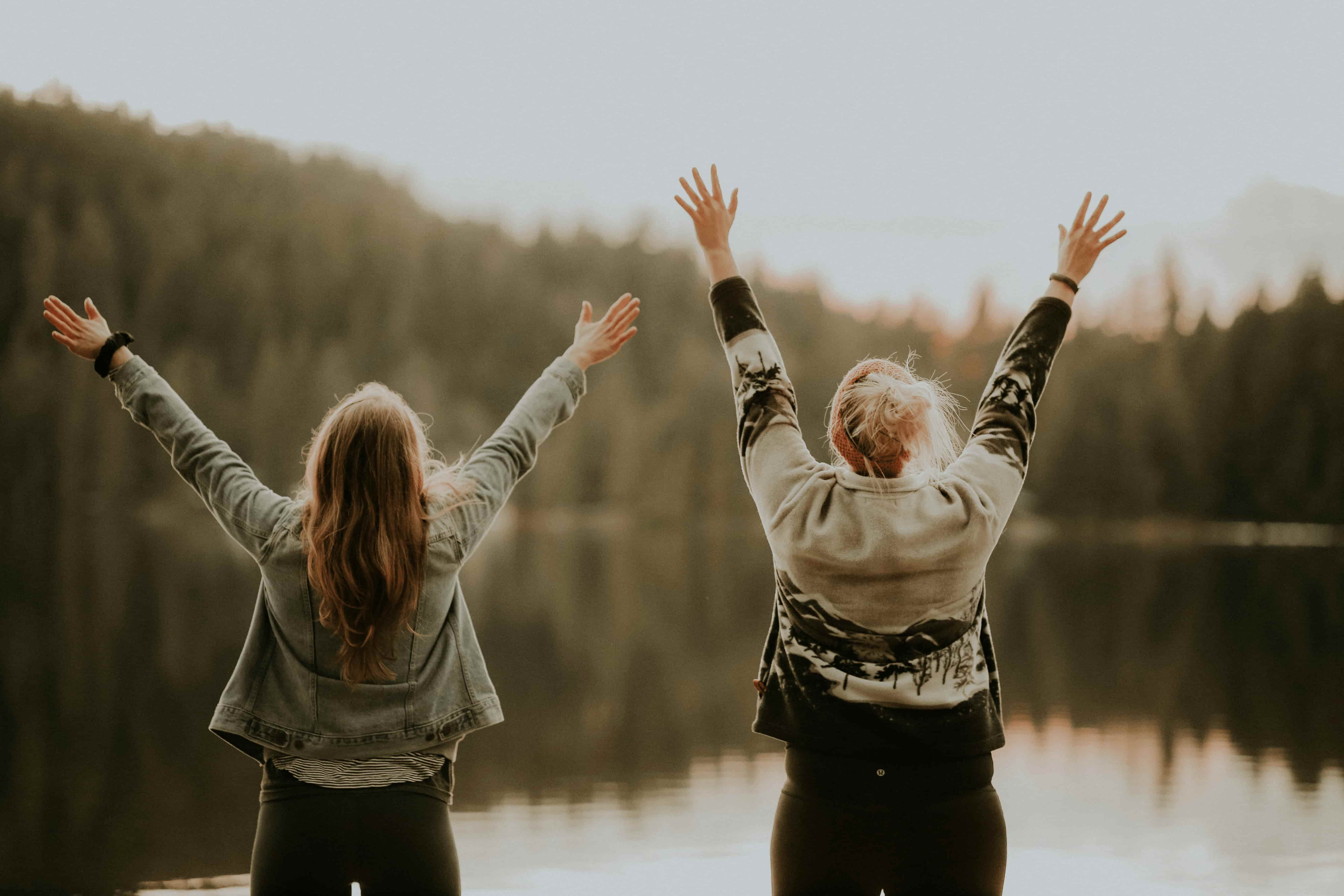 Two women standing with arms raised in celebration, facing a lake surrounded by forest.