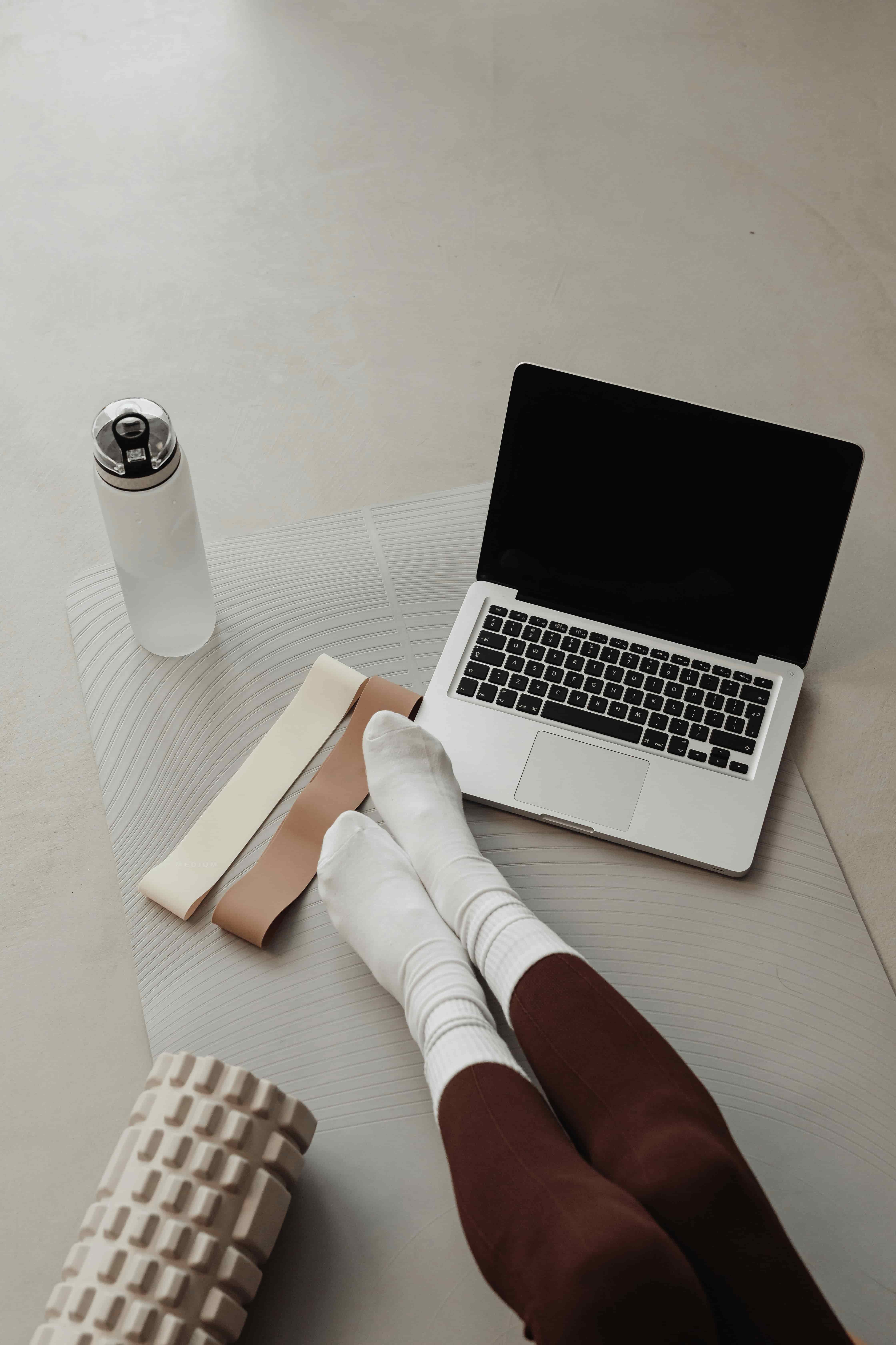 Person's legs in white socks resting near an open laptop, with a water bottle, resistance bands, and a foam roller on a yoga mat.