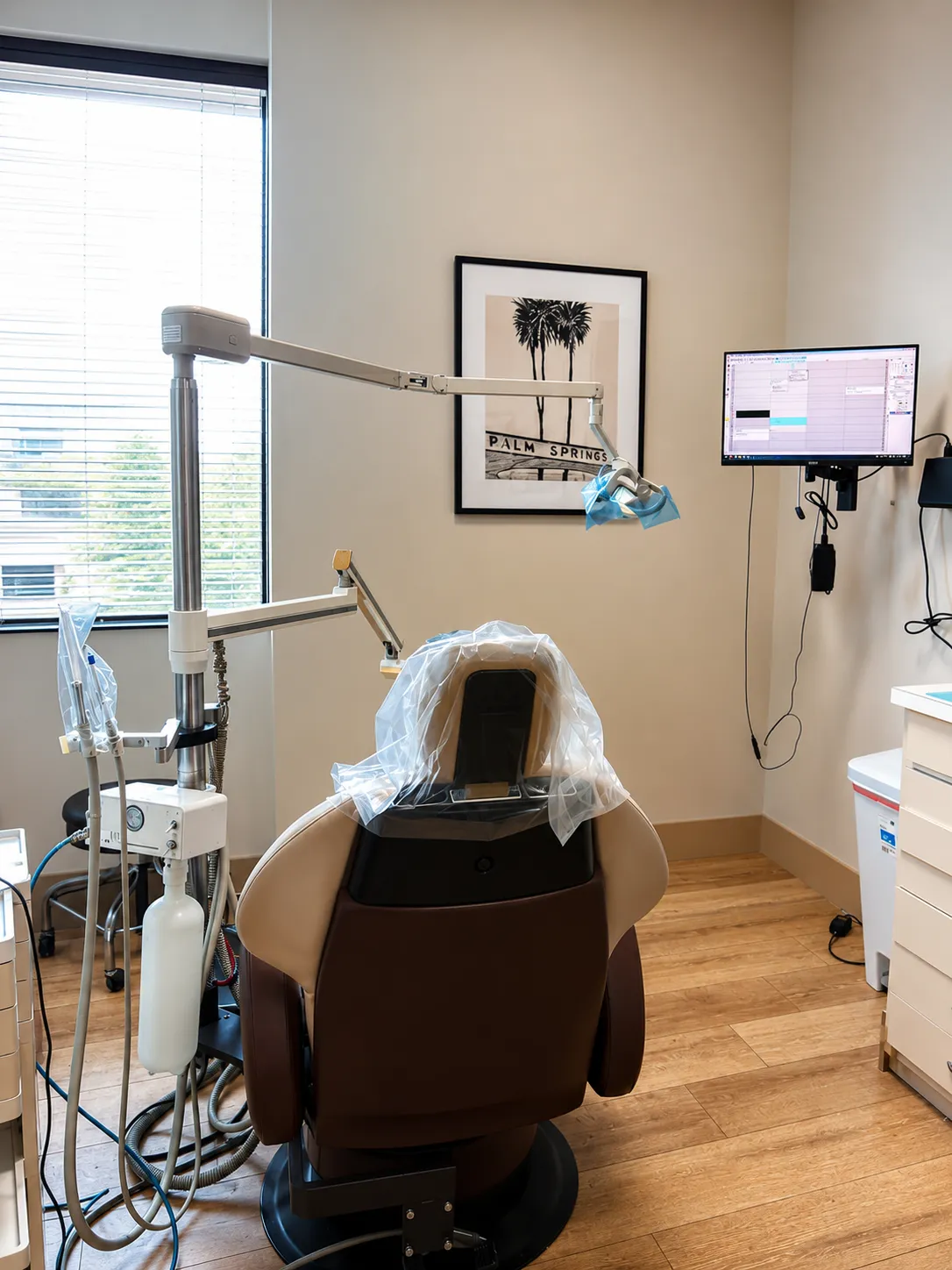 Dental chair facing a window with equipment, a Palm Springs photo, and a monitor on the wall.