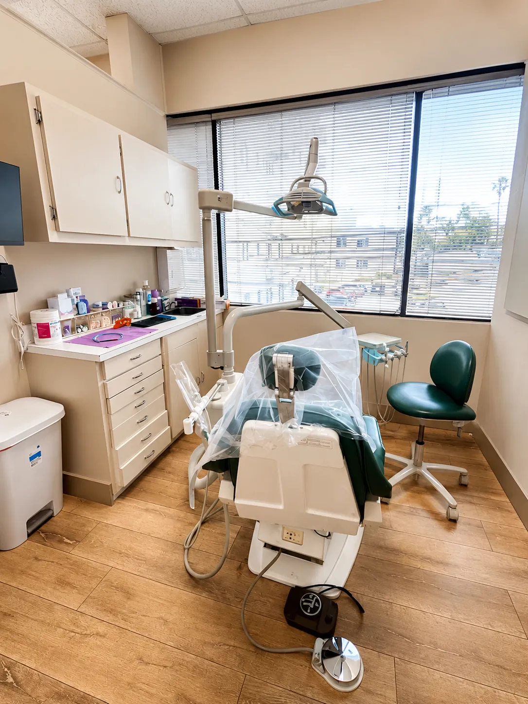 Dental office with a green dental chair, examination light, cabinets, and a window with blinds.