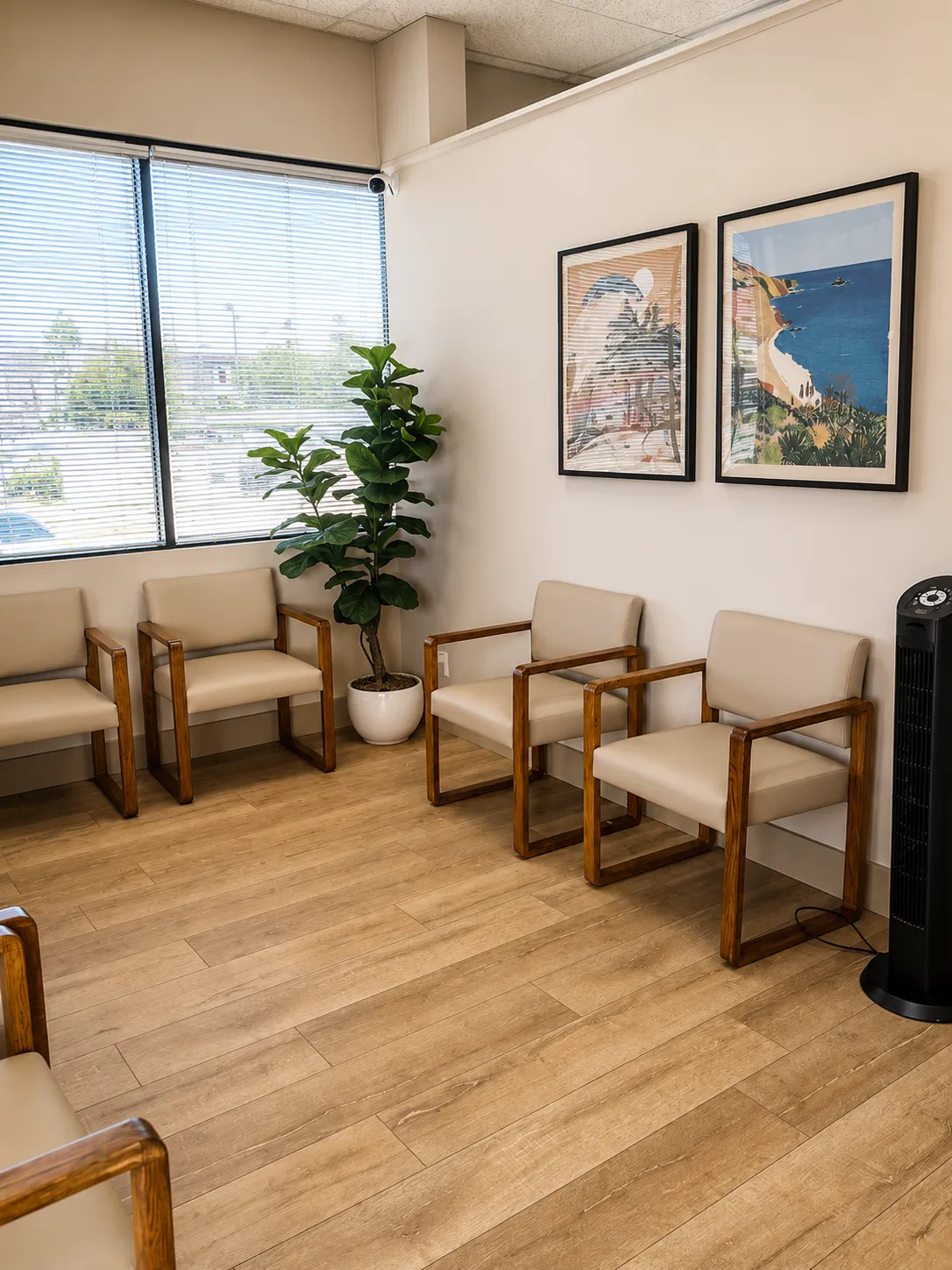 Waiting room with beige chairs, a tall green potted plant, two framed coastal paintings, and a black fan.