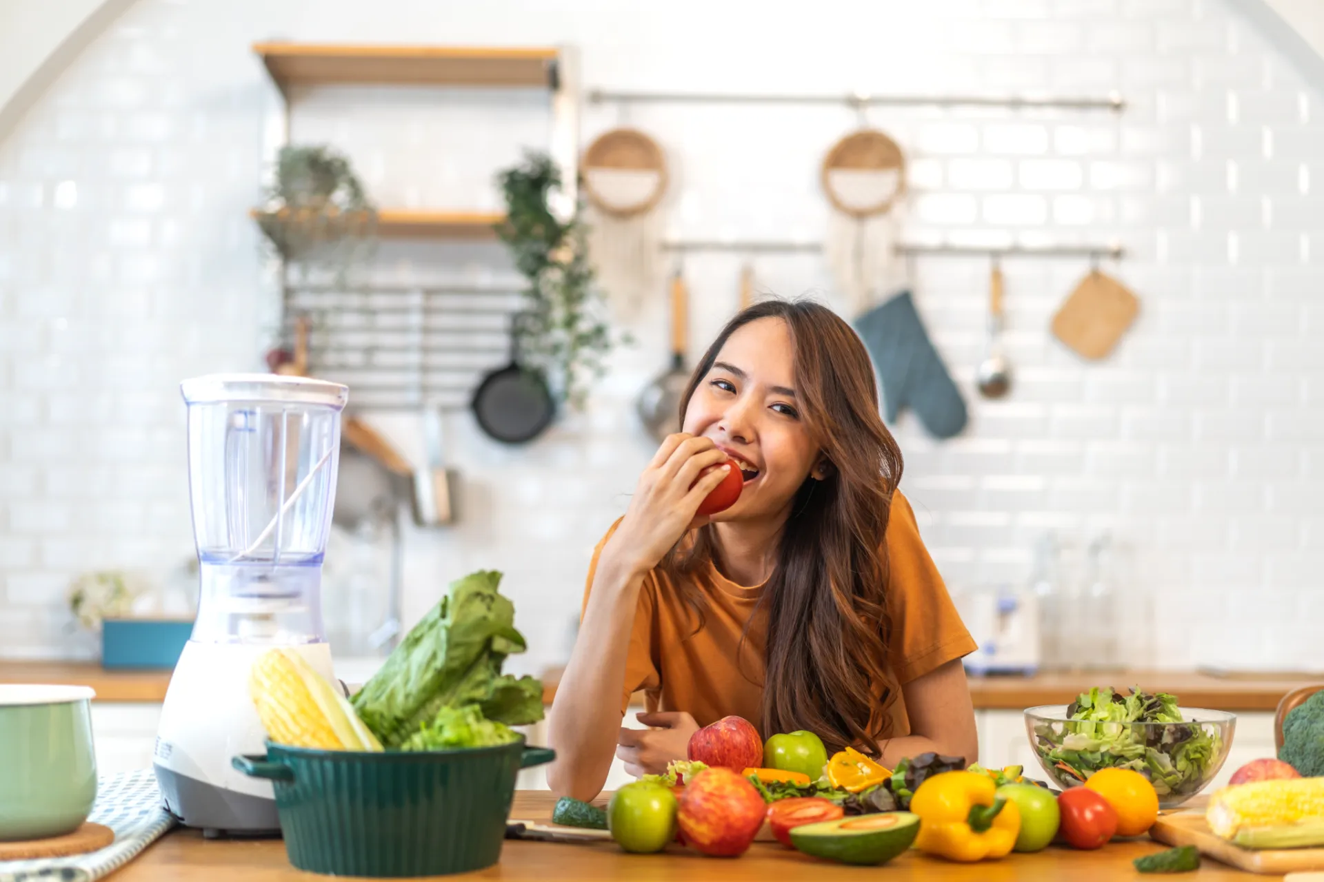 Young woman eating a tomato at a kitchen table with fresh vegetables and a blender nearby.