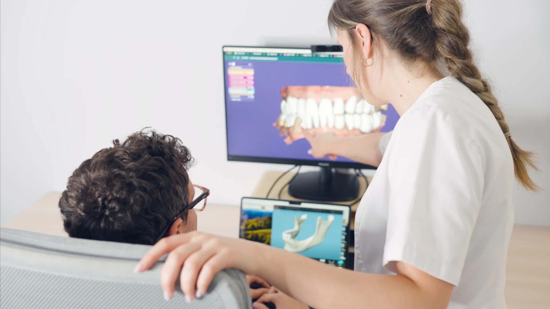 Dental professional showing a 3D teeth model on screen to a seated patient.