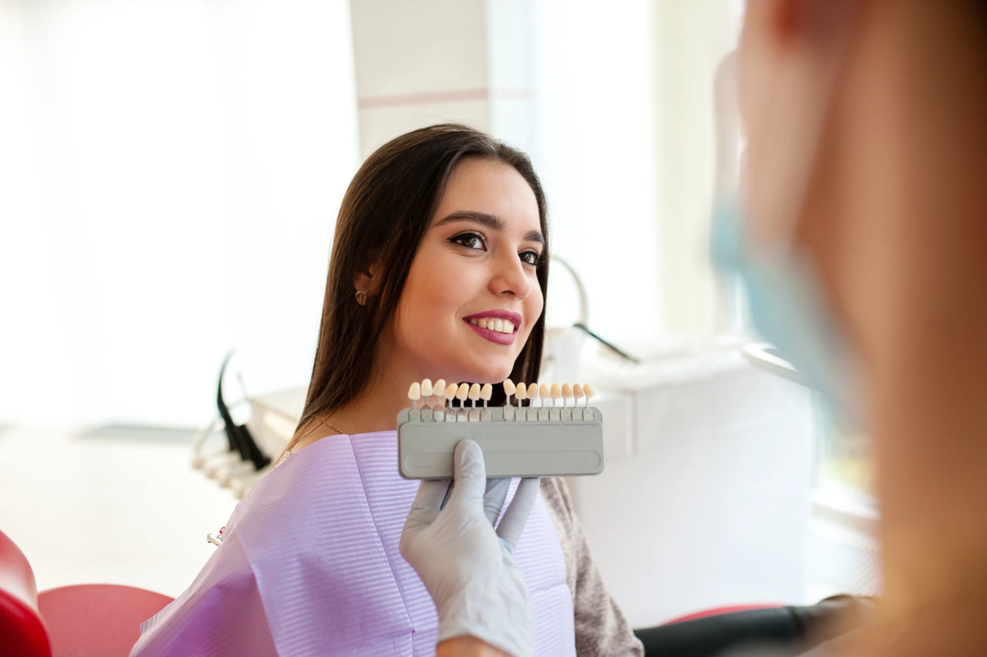 Dentist holding a tooth shade guide next to a smiling woman wearing a dental bib.