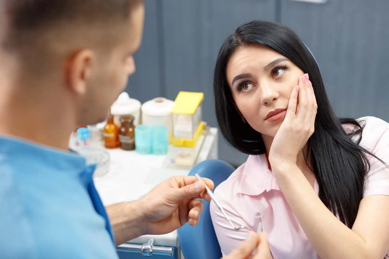 Woman in a dental chair holding her cheek while dentist shows her a dental tool.