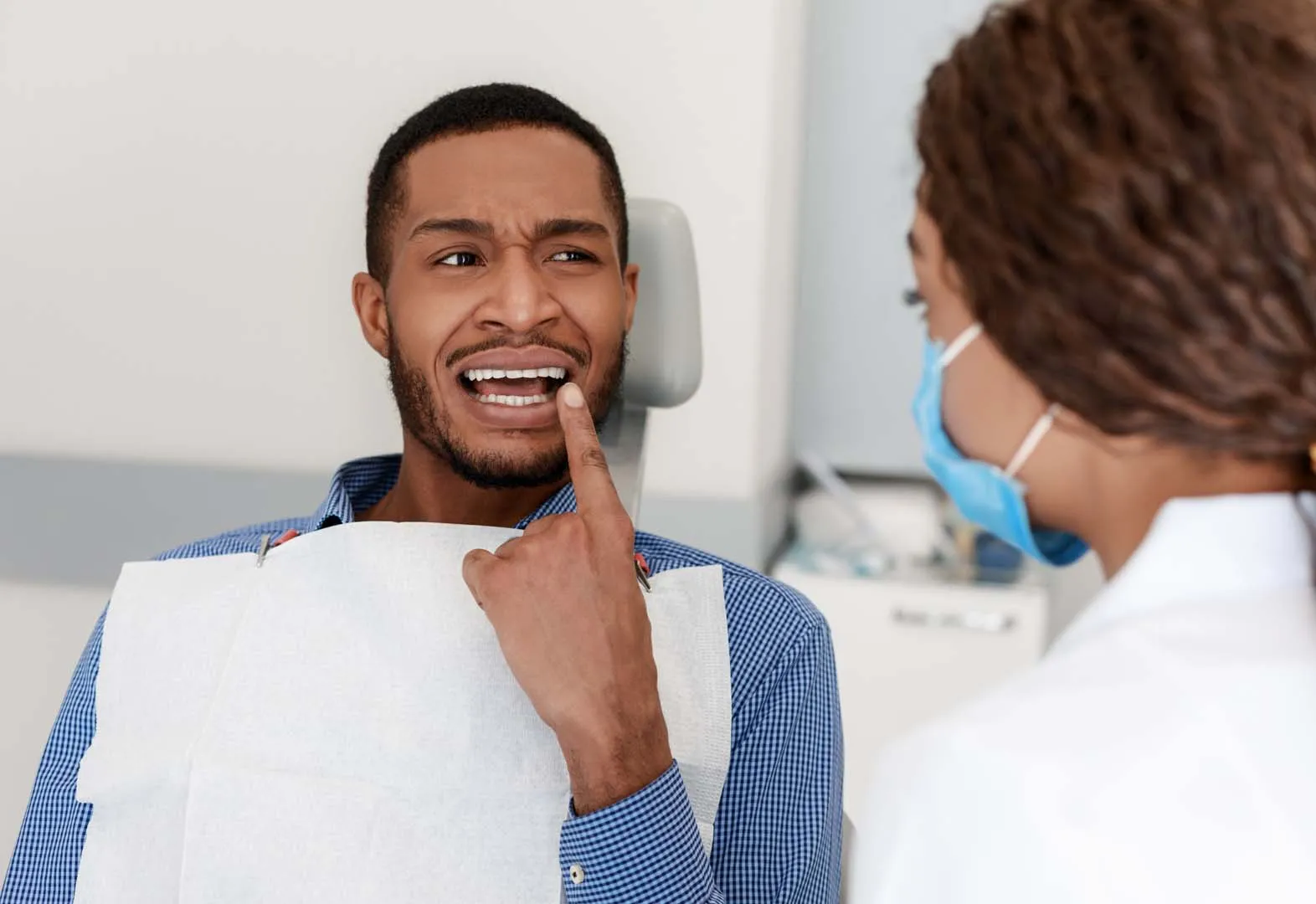 Man in dental chair pointing to his tooth while talking to masked dental professional.