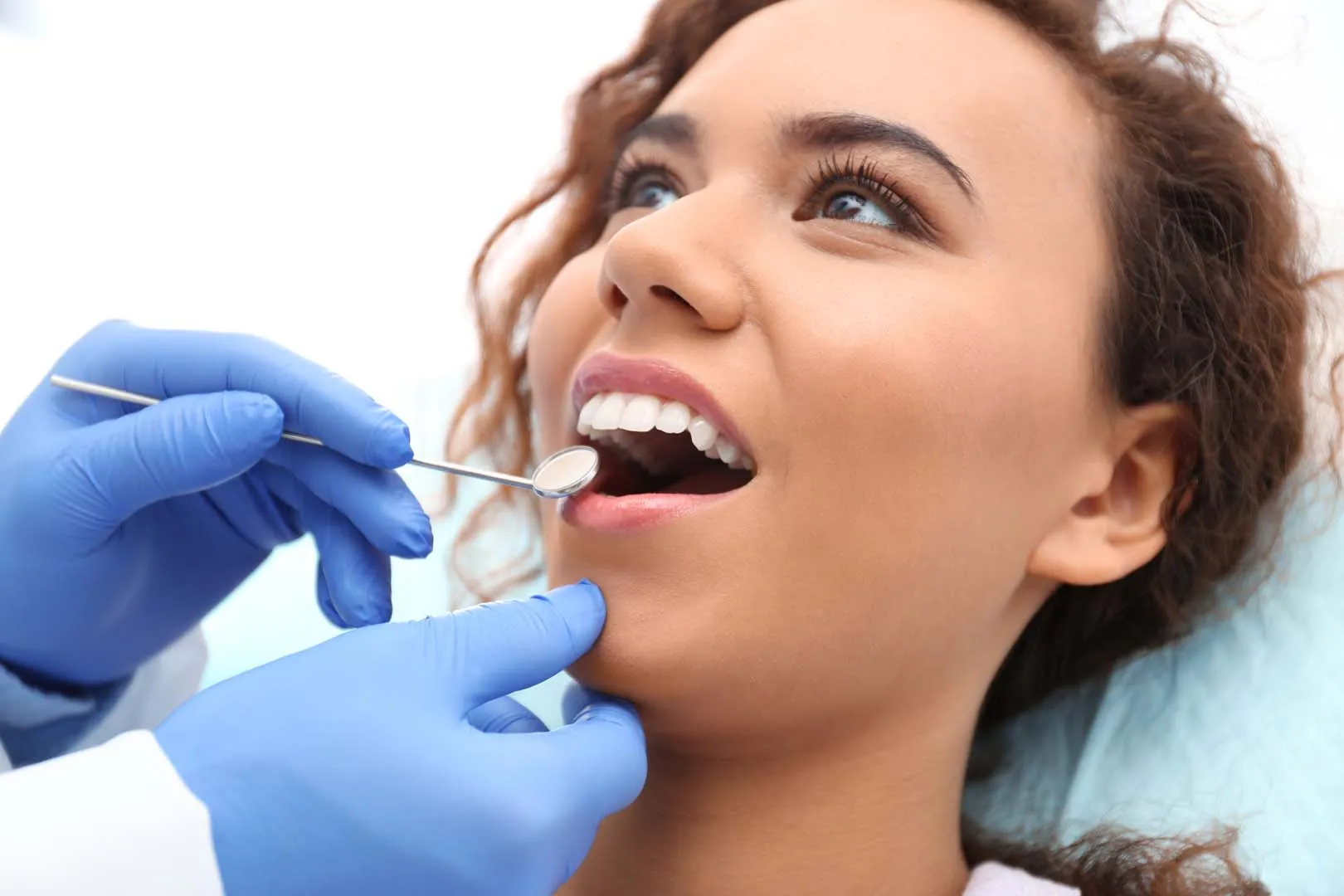 Dentist wearing blue gloves examining a smiling woman's teeth with a dental mirror.