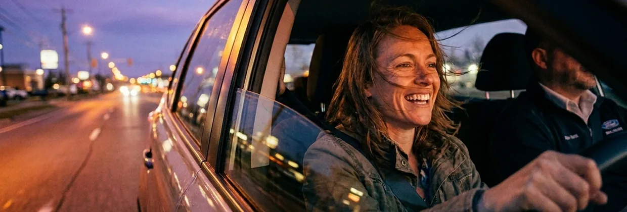 A smiling woman driving a car in twilight with her window rolled down.