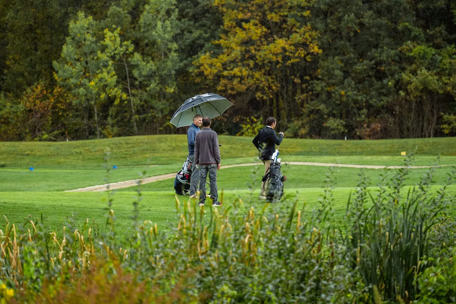 Drei Golfer mit Regenschirm und Golfbags auf dem Abschlag am Golfpark am Löwenhof, dahinter herbstlicher Wald
