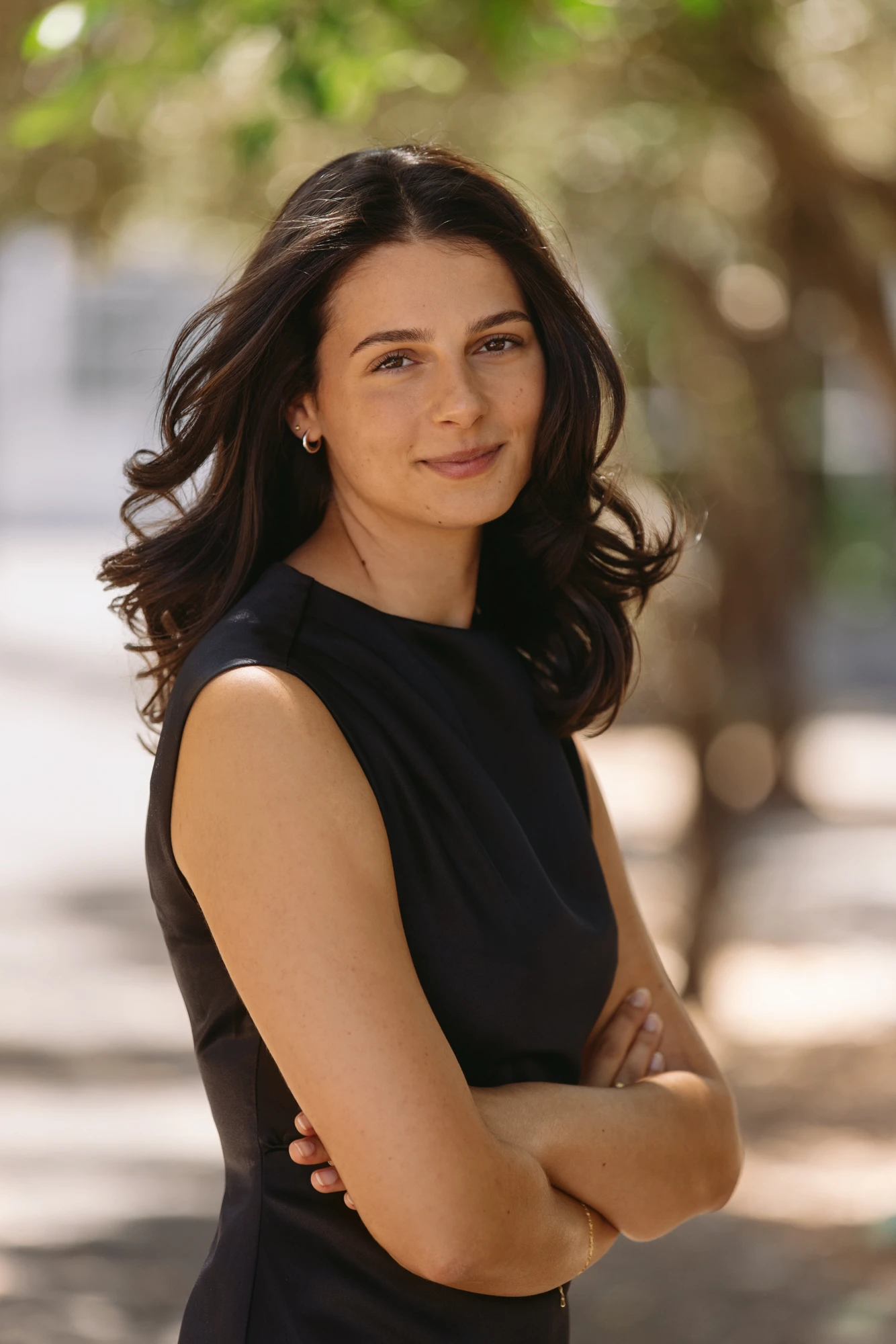 Smiling woman with dark hair in a sleeveless black top standing outdoors with arms crossed.