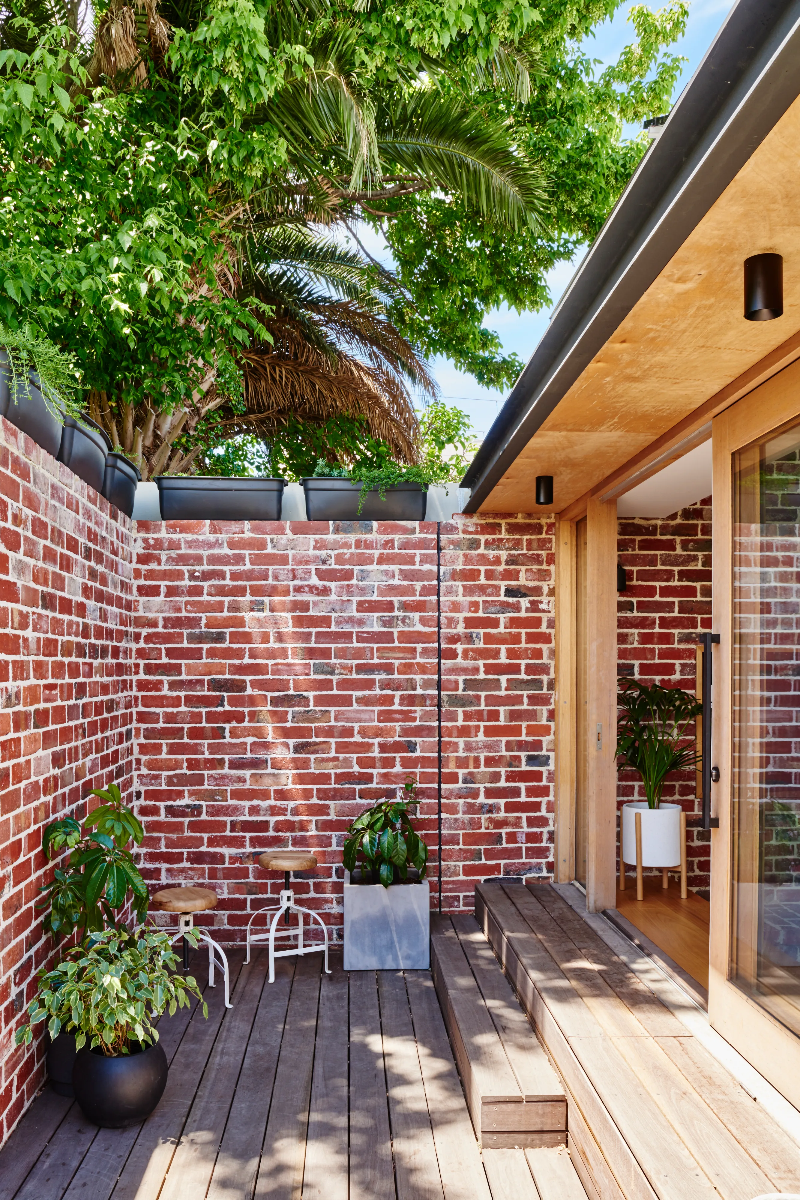 Small patio with wooden deck, brick walls, potted plants, stools, and sliding glass door entrance.