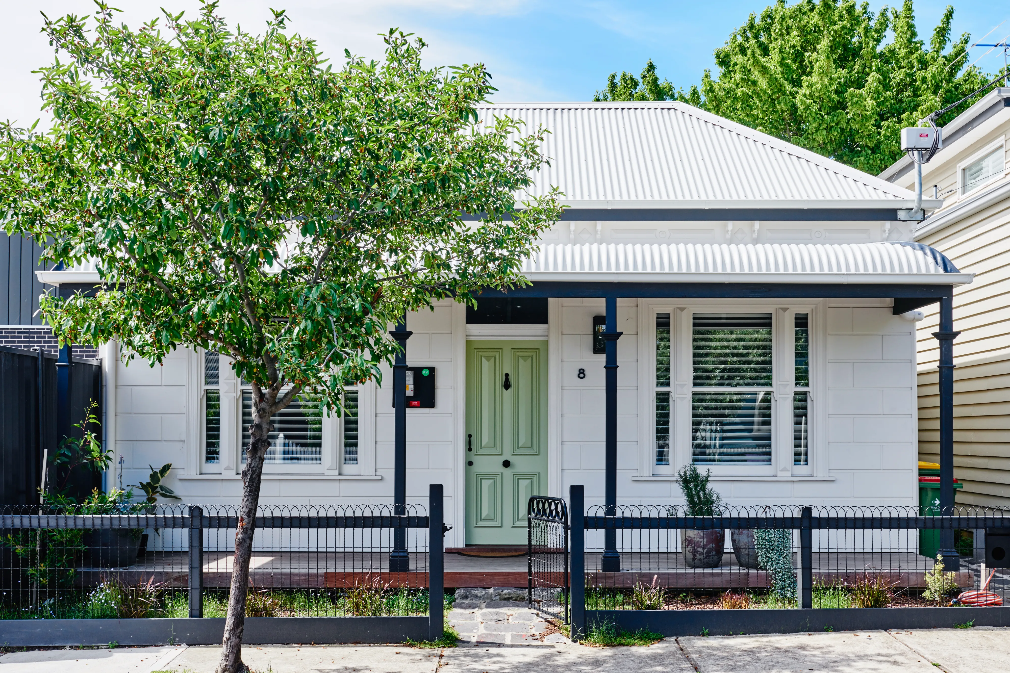 Small white house with green front door, black columns, and tree in front yard behind metal fence.