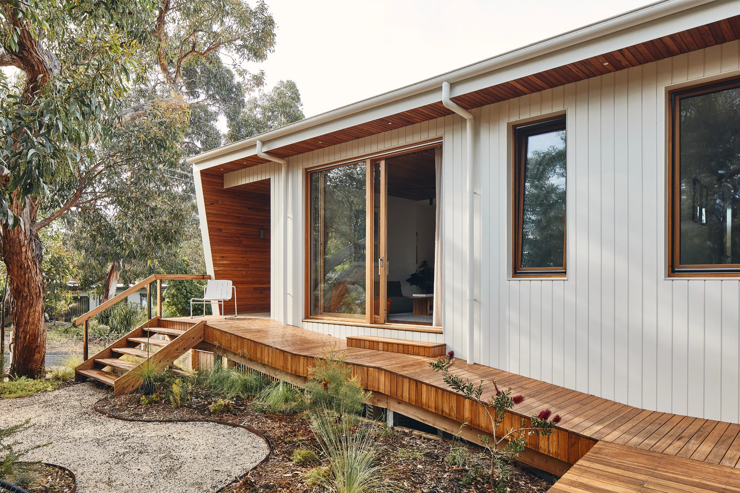 Modern house with wooden deck, sliding glass door, white siding, and surrounding greenery.