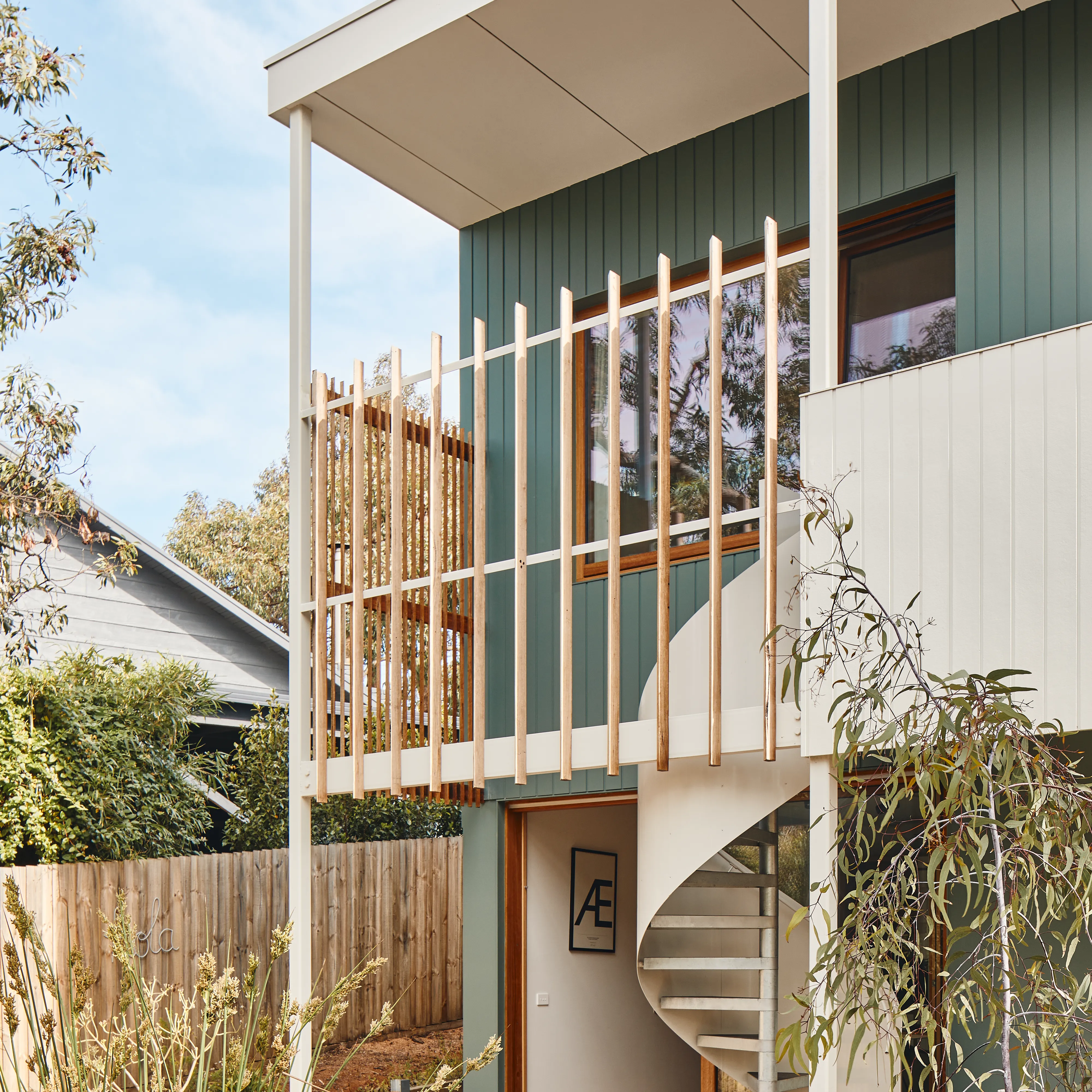 Modern two-story house with green and white vertical siding, large window, wooden vertical railing on balcony, and a white spiral staircase leading to the upper floor, set against a background of greenery and blue sky.