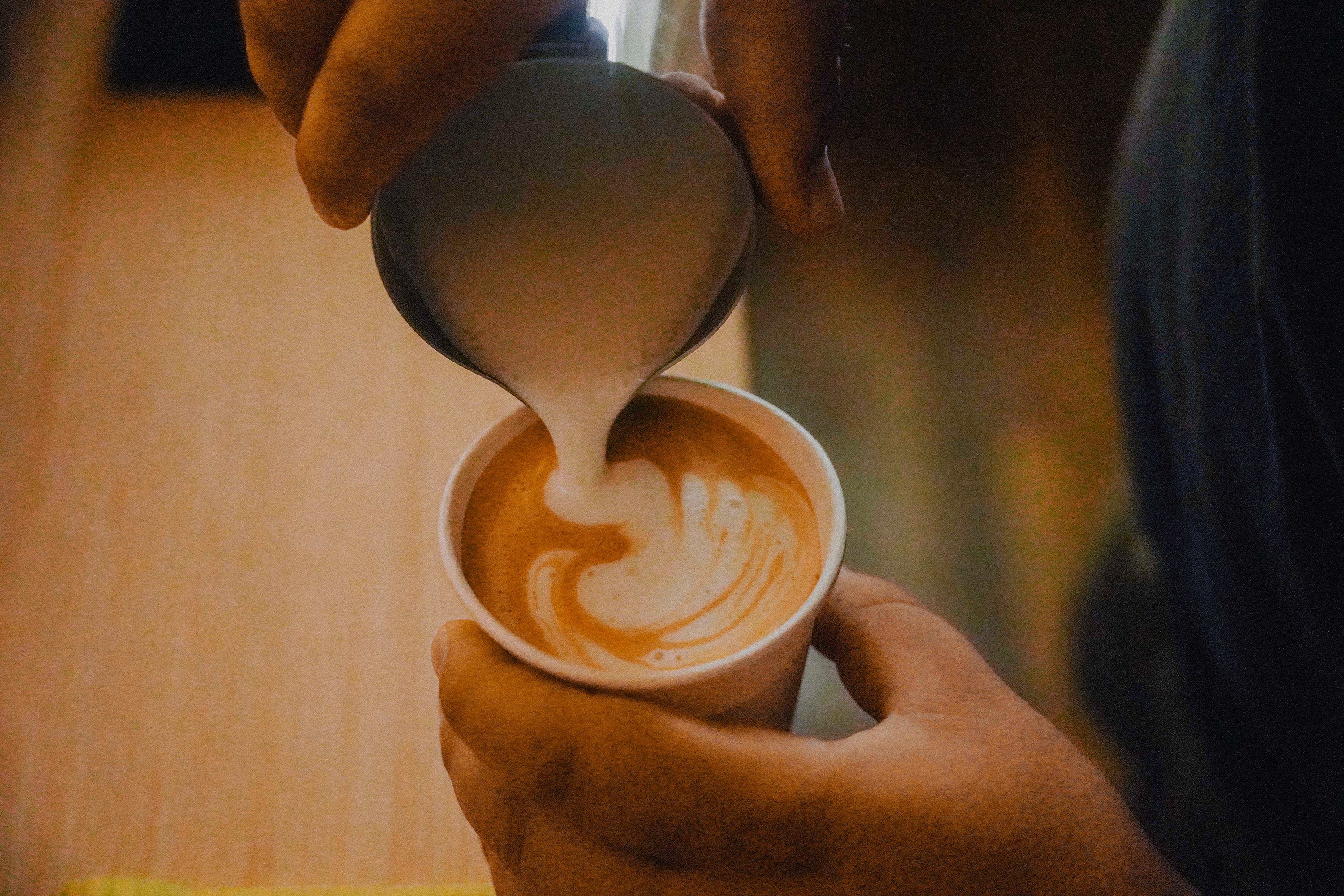 Close-up of hands pouring steamed milk into a cup of coffee to create latte art.