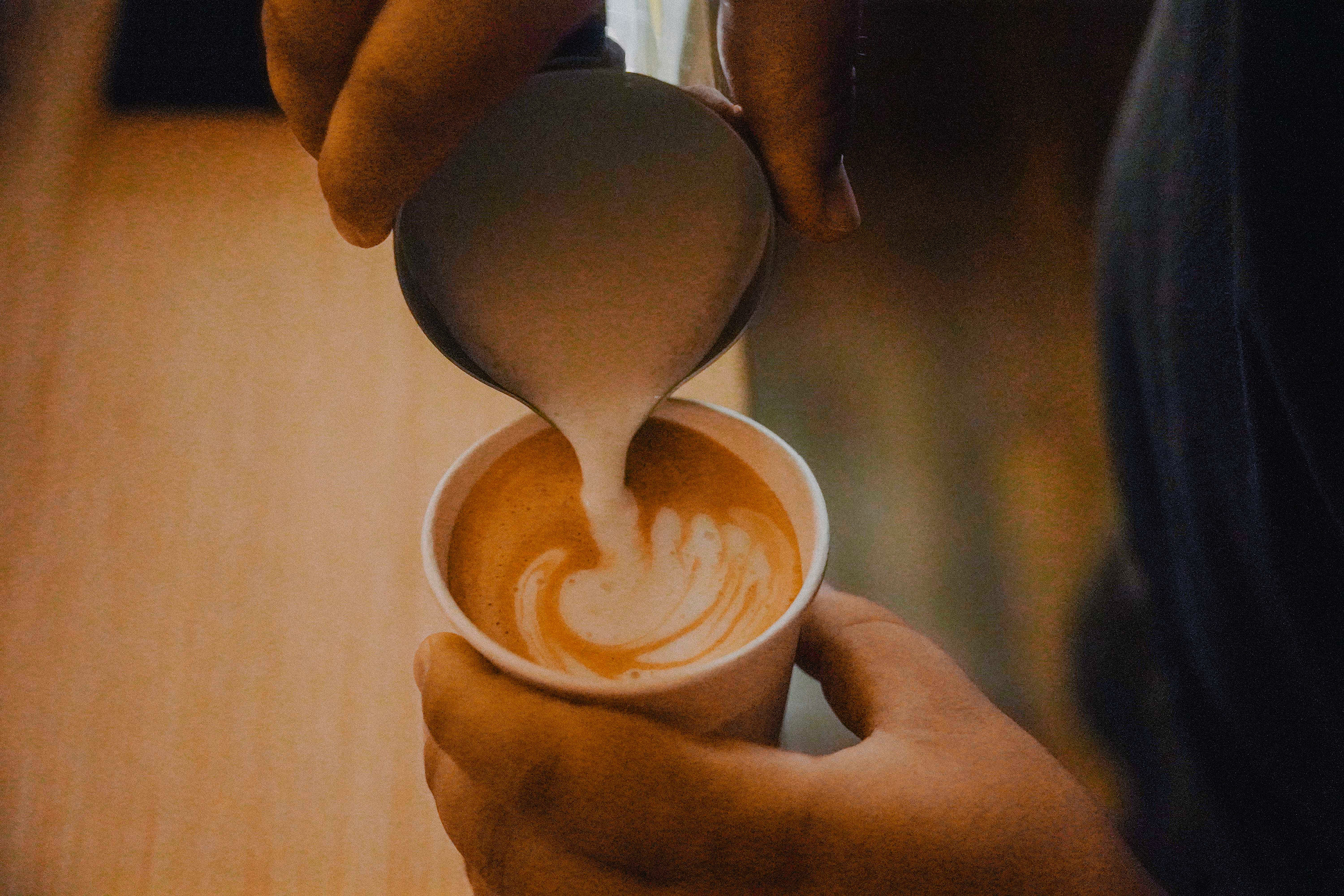 Person pouring steamed milk creating latte art in a cup of coffee held in hand.