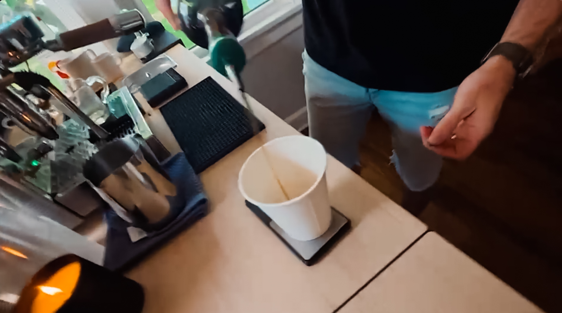 Person pouring a drink from a green bottle into a white cup on a scale on a counter.