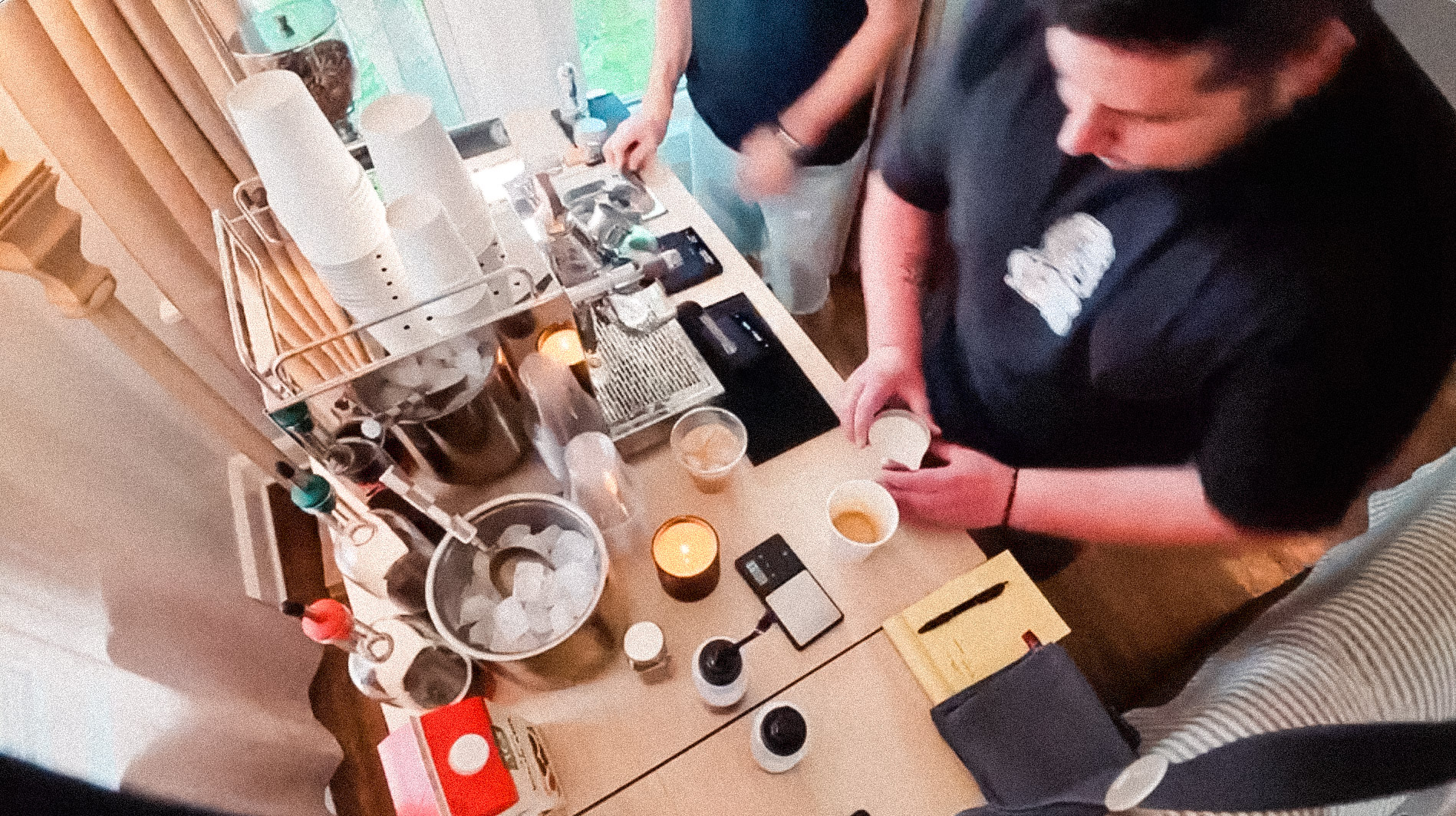 Top view of two men preparing drinks at a cluttered countertop with cups, ice, and coffee tools.
