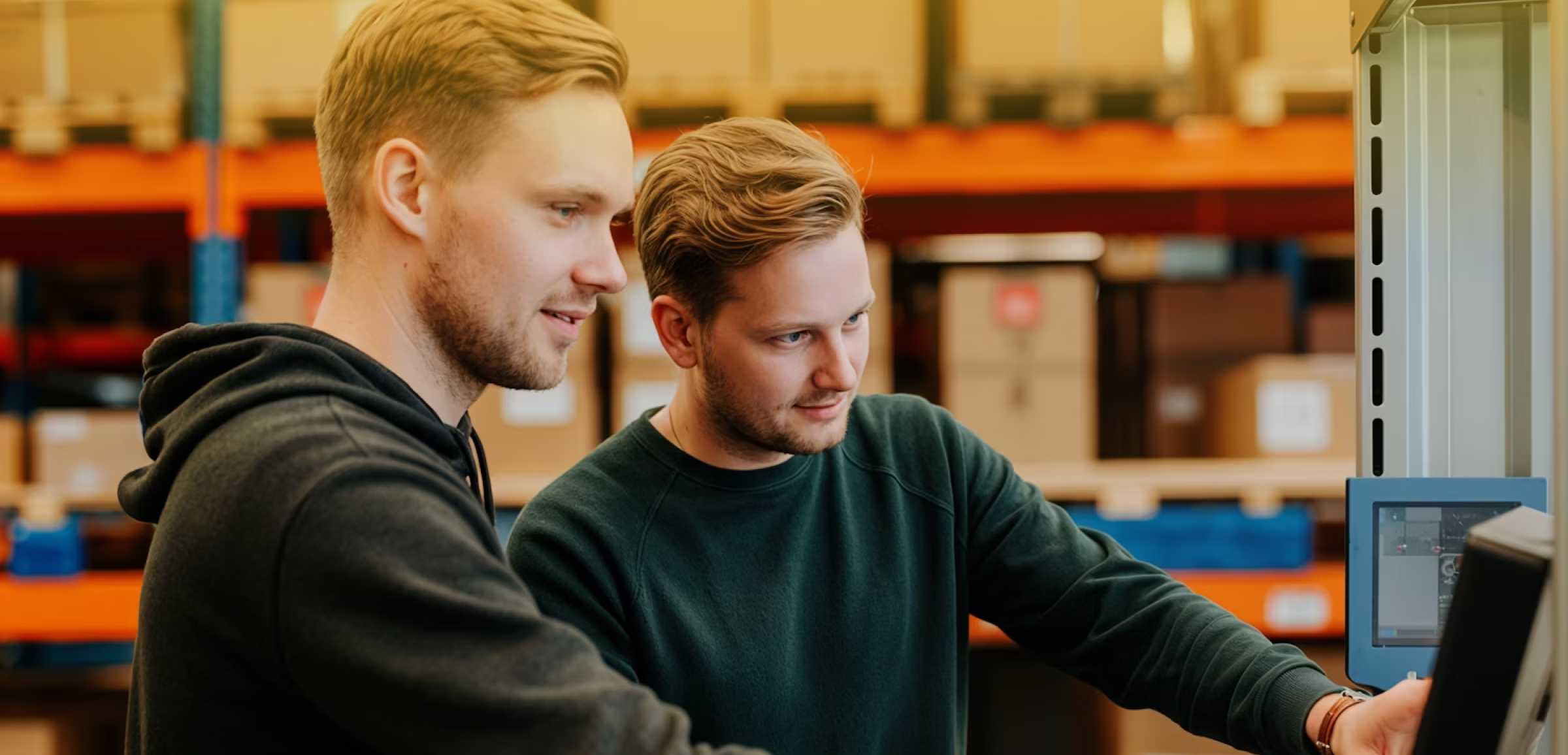 Two young men with short hair focus on a computer screen in a warehouse setting.