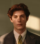 Young man with styled brown hair wearing a suit, white shirt, and yellow patterned tie, against a muted background.