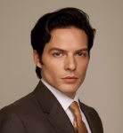 Young man with dark hair wearing a brown suit and light brown tie against a plain beige background.