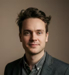 Headshot of a young man with dark hair, wearing a dark blazer and shirt, against a plain brown background.