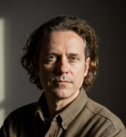Man with curly dark hair and a serious expression wearing a brown shirt in soft lighting.