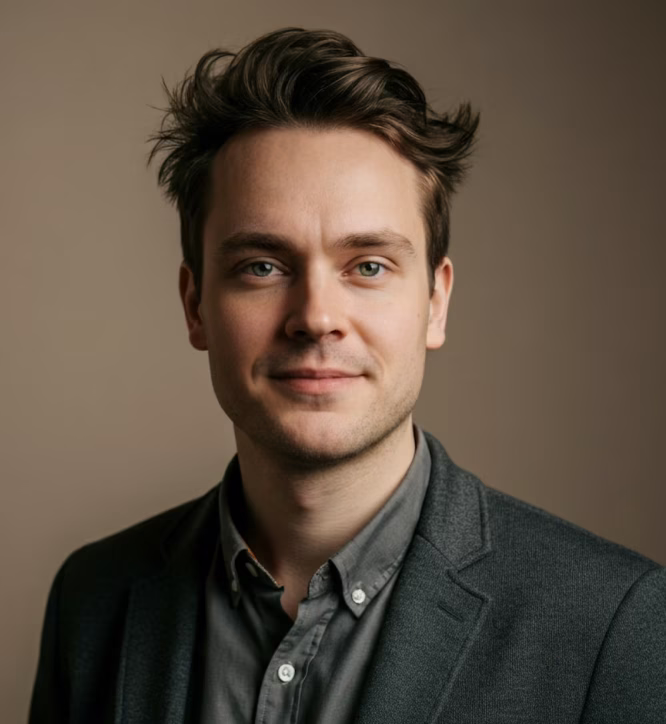 Portrait of a young man with short dark hair wearing a gray shirt and dark blazer against a plain beige background.