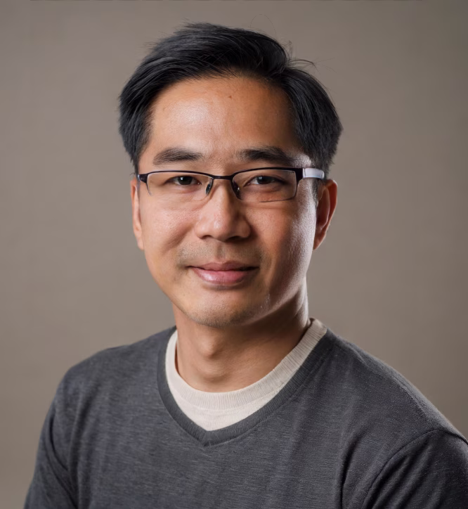 Portrait of a man with short black hair and glasses wearing a gray sweater over a white shirt, smiling softly against a neutral background.
