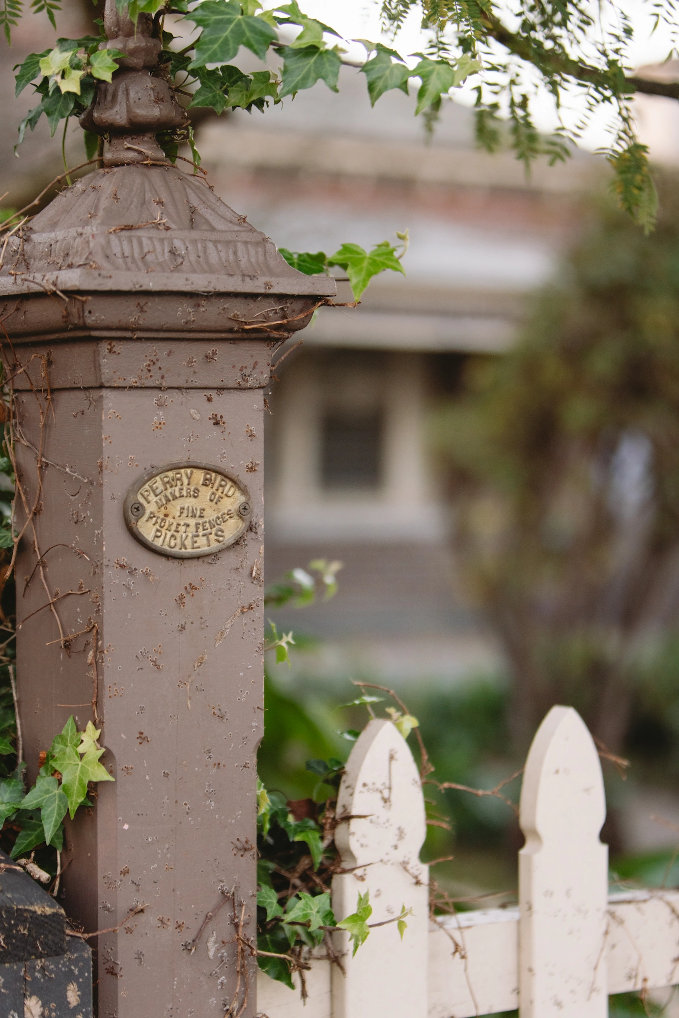 Close-up of a weathered cast-iron gatepost with a brass nameplate, beside a cream picket fence in a Hawthorn garden.